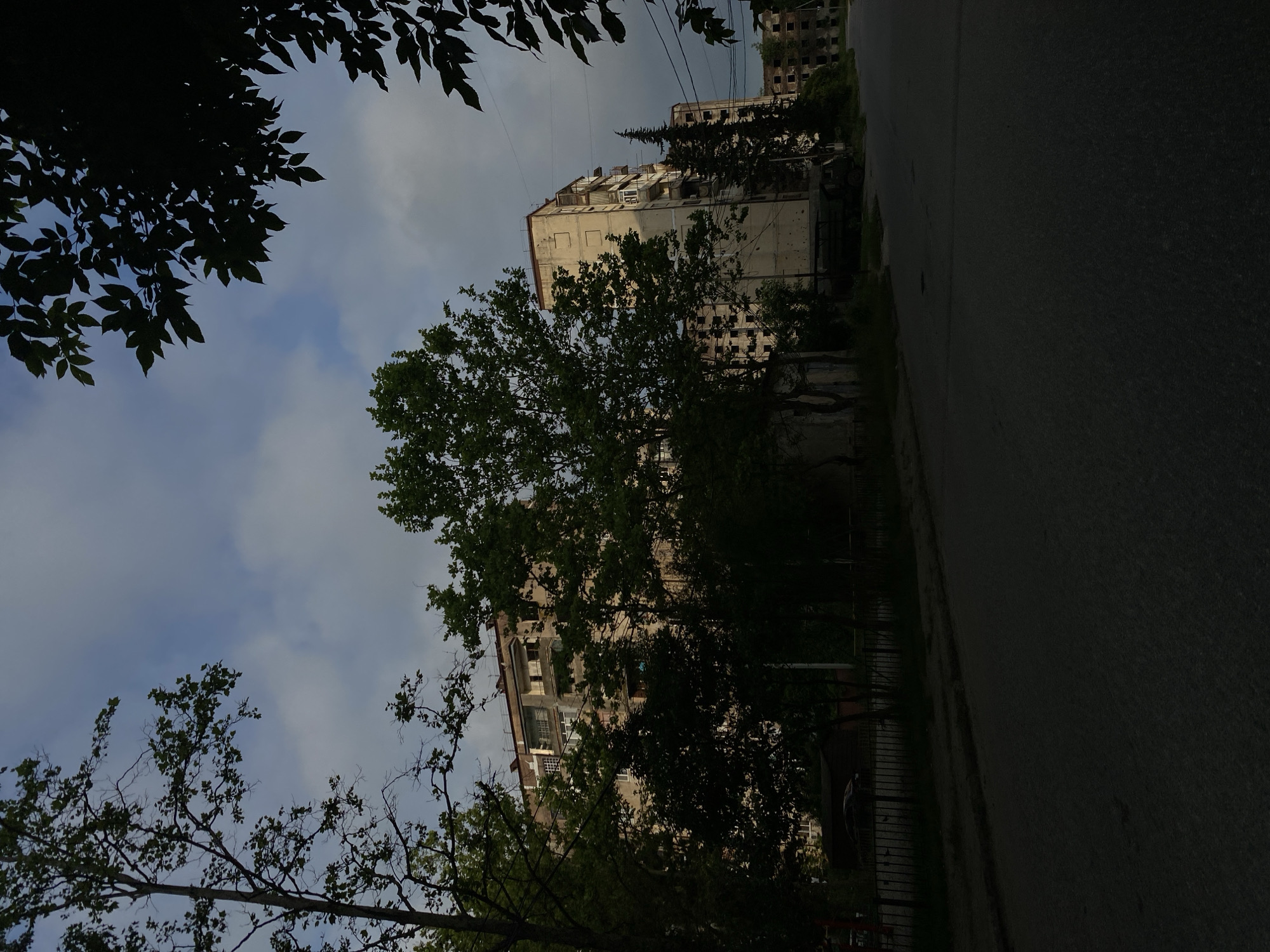 A street view shows apartment buildings on the right side of the road. The buildings are multi-story with balconies and windows. Trees are in the foreground and scattered around the buildings. The sky is cloudy. A paved road is in the foreground, with a sidewalk and fence to the left of the road. There are no people visible in the image.