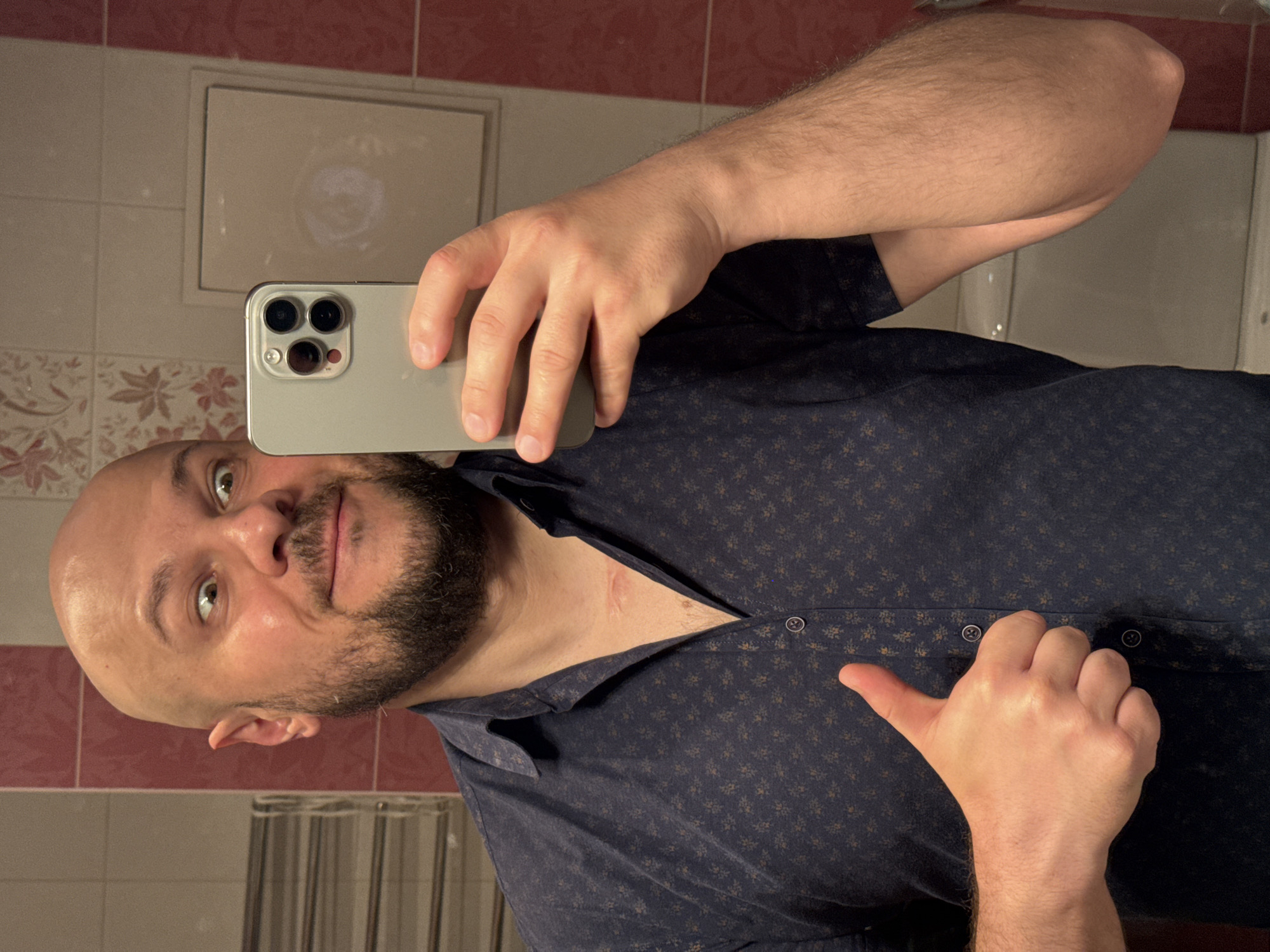 The media file is a selfie taken in a bathroom. The subject is a bald man with a beard, wearing a dark blue patterned shirt. He is holding a smartphone in his left hand, capturing his reflection in a mirror. His right hand is extended, giving a thumbs-up gesture. The background shows tiled walls, with the top portion being a reddish-pink, and the lower portion lighter with a floral pattern. A small light-colored cabinet is visible on the wall. The lighting suggests it is likely daytime, possibly with artificial bathroom lighting. The setting appears to be a domestic bathroom, possibly in Almaty, Kazakhstan, as hinted by the location context. The man's expression is neutral, and the thumbs-up gesture suggests a positive sentiment. No visible text is present in the image.