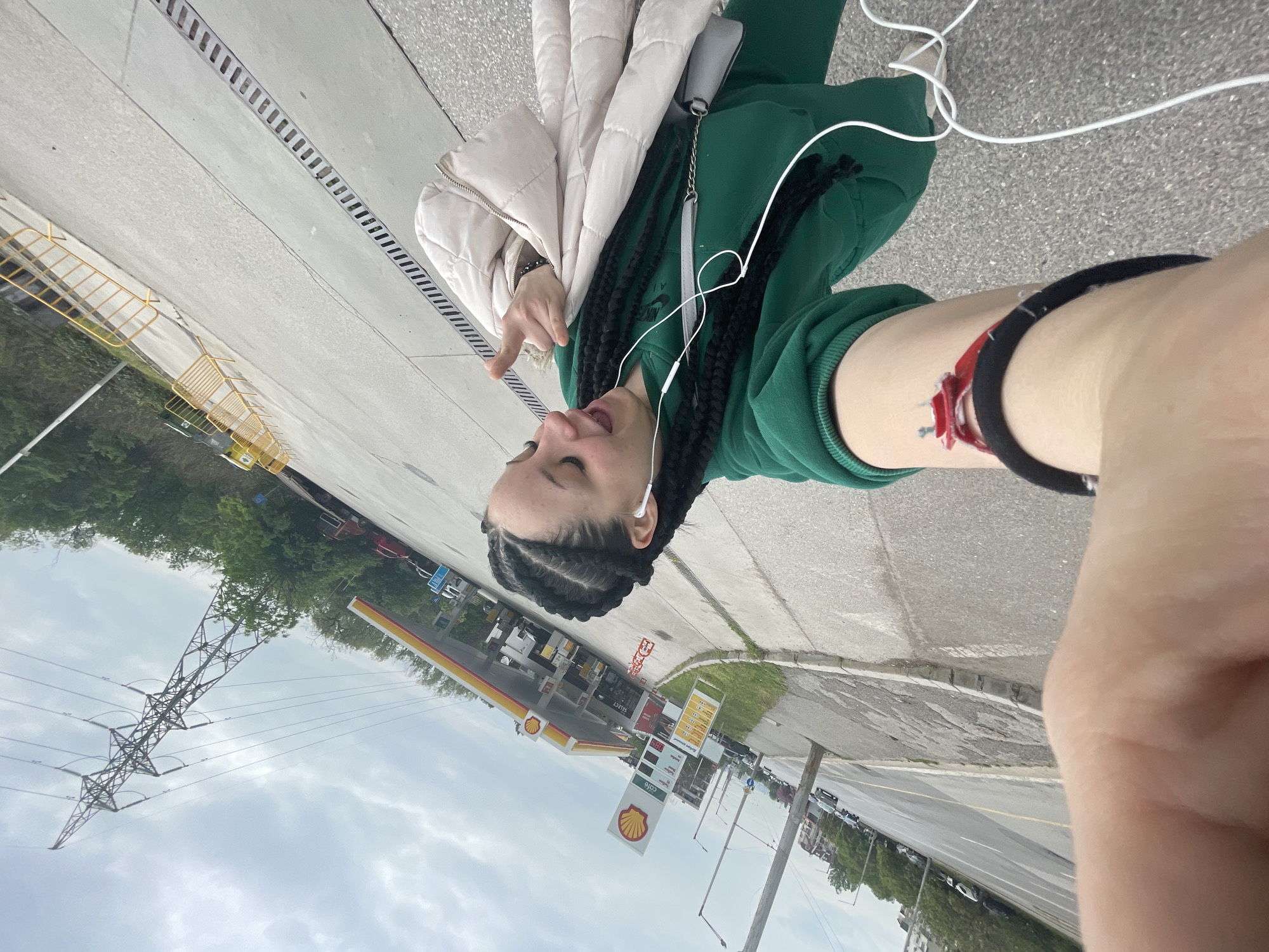 A selfie photo shows a person with dark braided hair, wearing a green tracksuit and a light-colored puffer jacket, standing next to a Shell gas station in Sofia, Bulgaria. The person has earbuds in their ears and is making a hand gesture. An electrical tower is visible in the background. The photo seems to have been taken from a low angle, possibly with a phone camera.