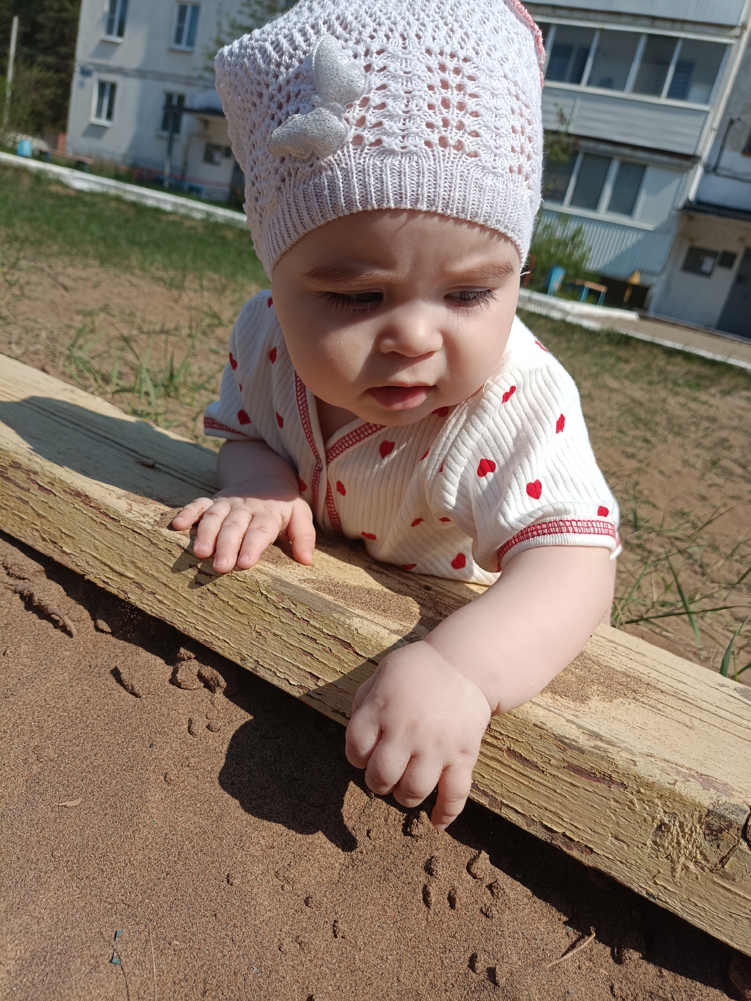 A close-up, eye-level shot shows a baby leaning over a weathered wooden structure, seemingly in a sandbox, located outside in Noviy, Russia. The baby wears a white crocheted hat with a decorative butterfly on it, and a white shirt with small red hearts. The baby's right hand is reaching toward the sand, as if exploring it. The backdrop features a light-colored building with windows and possibly some green space or grass. The lighting is bright, suggesting it is daytime.