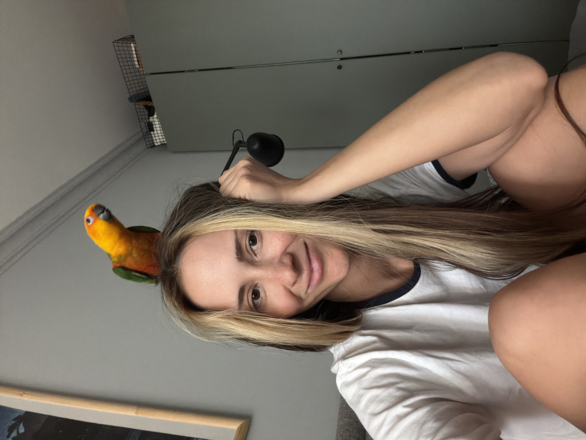 A close-up selfie of a young woman with blonde and brown hair is taken indoors, likely in an apartment. She is wearing a white t-shirt with a dark blue trim. Her right arm is bent, and she is holding a black object, possibly a microphone or a light, near her ear. A brightly colored parrot, with yellow, orange, and green feathers, is perched on top of her head. The background shows a gray wall, a closed closet with two doors, and a wire basket on top of the closet containing some items. The overall impression is of a casual, indoor setting. The provided metadata does not contain information about the location in the city of Khimki, Russia.