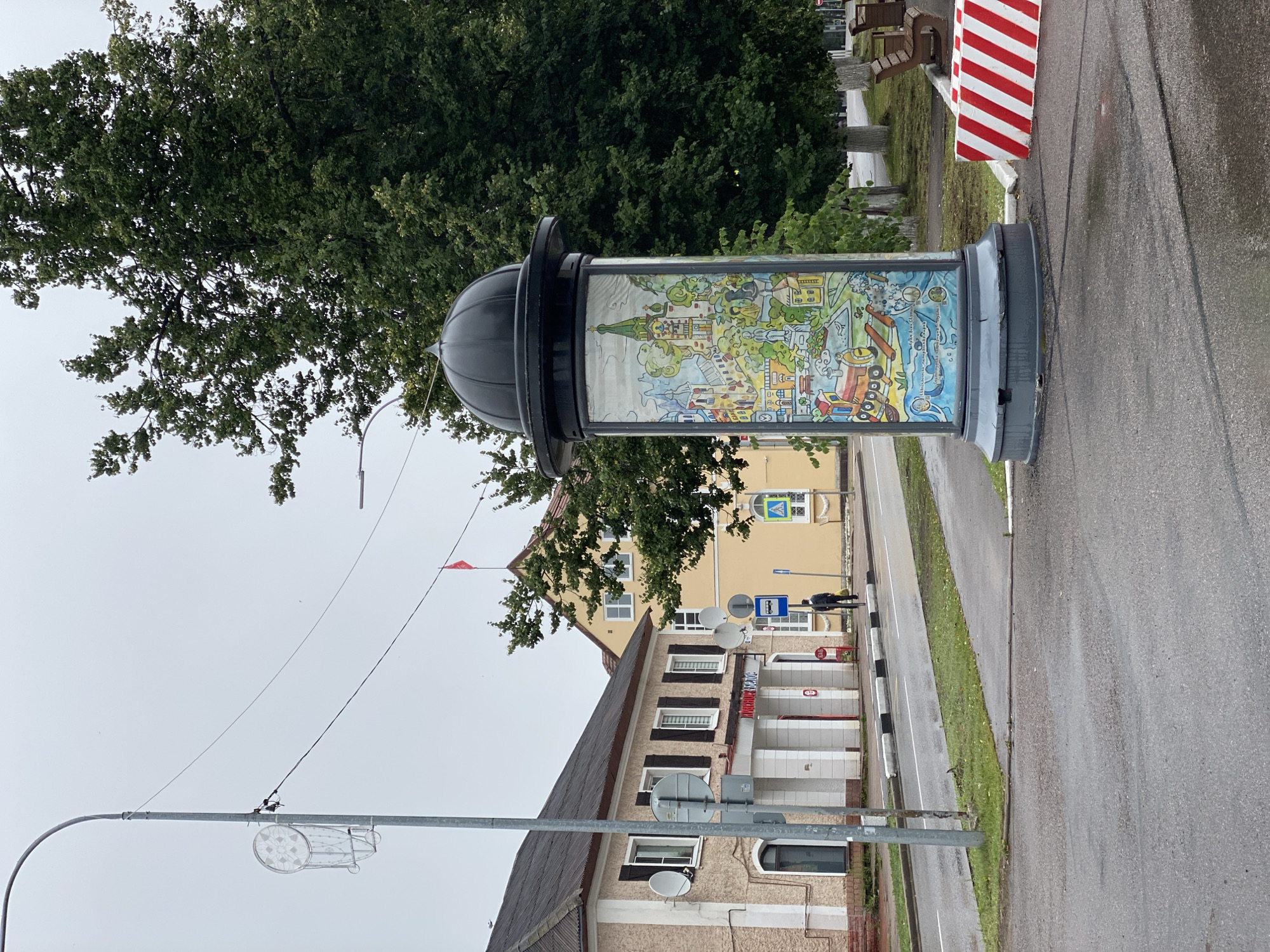 The image shows a street scene in Priozersk, Russia, on an overcast day. In the foreground, a decorative pillar stands on a sidewalk. The pillar is cylindrical and topped with a dark dome. Its sides are covered with a colorful mural depicting various buildings, a train, and water. To the right of the pillar, a red and white striped barrier is visible.
On the street, a gray asphalt road is wet, suggesting recent rain. A sidewalk runs alongside the road, with a pedestrian crossing marked by black and white stripes. A streetlamp with a circular decorative element stands on the left side of the image.
Behind the pillar and sidewalk, a light-colored building with brown shutters and arched windows is visible. A sign on this building appears to be in Russian. Further down the street, a yellow building with a red roof and a red flag on top is also visible.
A single person, dressed in dark clothing and carrying a bag, is walking on the sidewalk in the middle ground, heading away from the camera. Several trees with dense green foliage are present, framing the scene and casting shadows. Two wooden benches are placed on the grass to the right of the pillar, near the barrier. Satellite dishes are mounted on the side of the light-colored building.