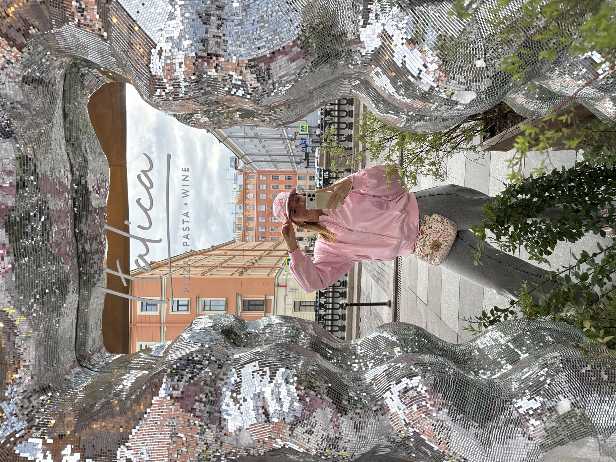 A woman in a pink jacket and cap stands next to a mirrored sculpture on a city street, holding a phone in her hand and possibly taking a photo or selfie. The sculpture is composed of small, reflective squares arranged in a wavy or undulating pattern. Through the opening in the sculpture, the facade of a building with multiple windows can be seen, along with a street sign. A sign that reads "Tatica - PASTA WINE" is visible. The location is in Saint Petersburg, Russia.