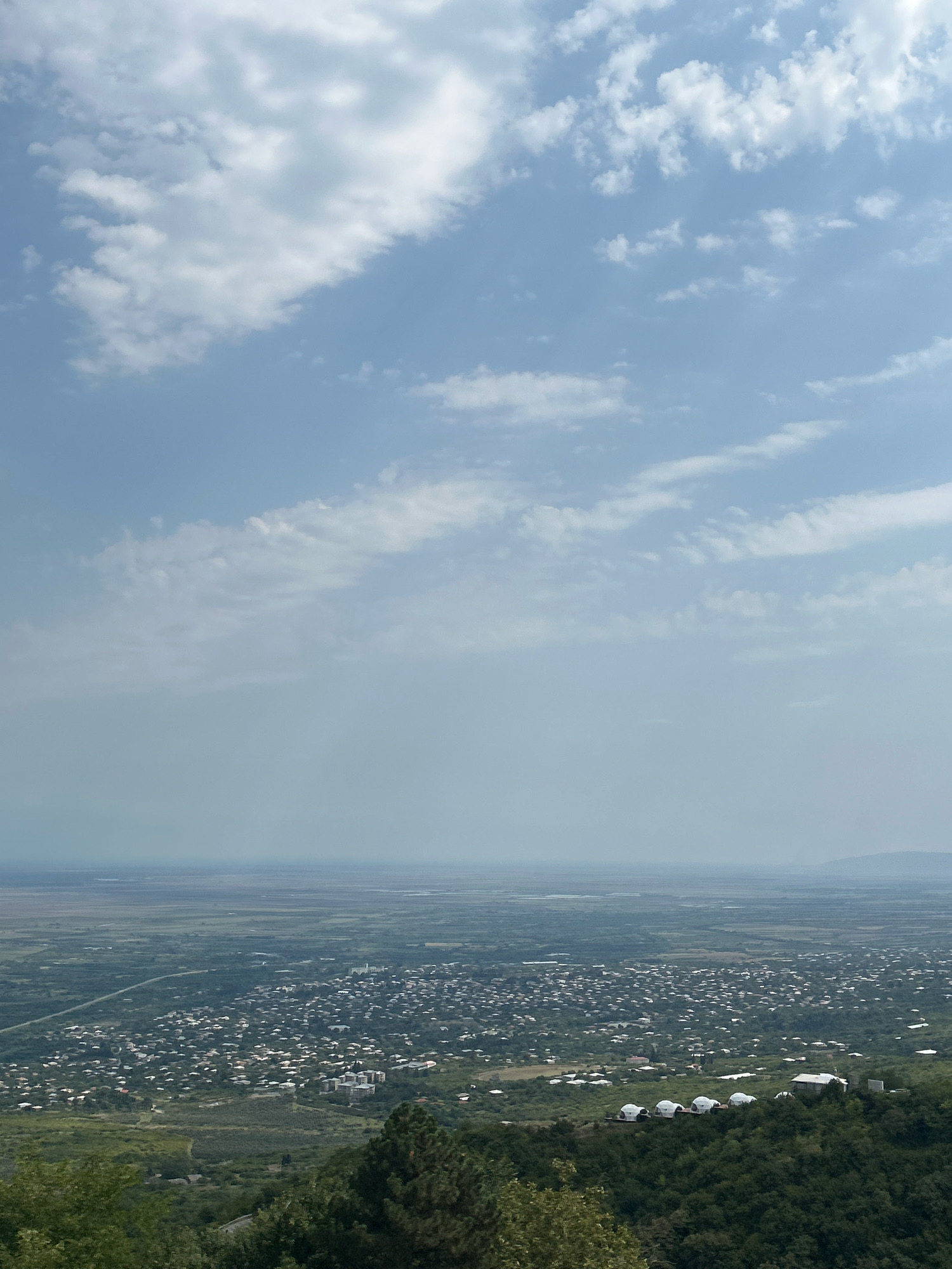 The image depicts a wide, elevated view of the city of Sighnaghi, Georgia. In the foreground, lush green trees dominate the lower portion of the frame. Beyond the trees, the city sprawls across a hillside, characterized by a dense collection of houses with red-tiled roofs. In the mid-ground, a cluster of dome-shaped structures, possibly accommodations, are visible on a lower ridge. The background reveals a vast, flat landscape that stretches to the horizon, where it meets a hazy, pale blue sky dotted with scattered white clouds. The overall scene conveys a sense of peaceful, expansive rural scenery.