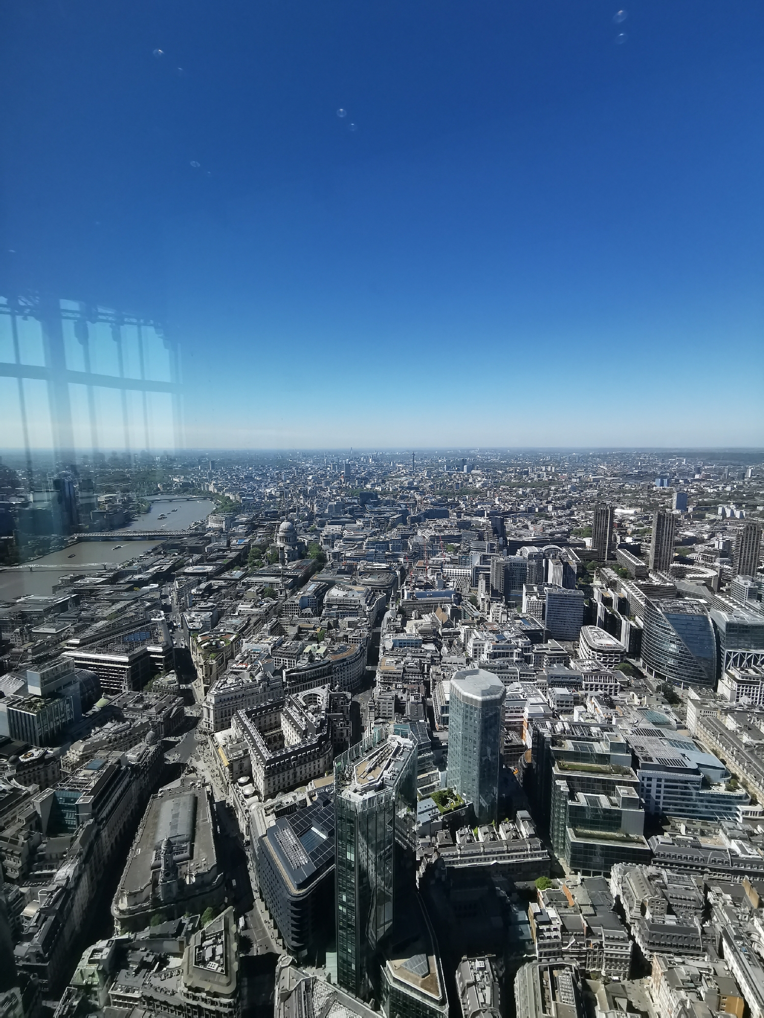 This image captures a sweeping aerial view of a vast, densely packed urban landscape, characteristic of London, United Kingdom. The photograph is taken on a clear, bright day, likely midday, with a deep blue, cloudless sky dominating the upper portion of the frame.
The city sprawls as far as the eye can see, featuring a complex tapestry of buildings that blend historical architecture with modern skyscrapers. The River Thames snakes through the left side of the scene, marked by several bridges and small boats. Prominently visible on the river's right bank is the distinctive dome of St. Paul's Cathedral, a classic London landmark.
Numerous contemporary glass and steel buildings punctuate the cityscape, including a notable, somewhat hexagonal or octagonal-shaped skyscraper in the lower-middle foreground. Other modern structures with geometric designs and rooftop gardens are interspersed among older, more traditional buildings with varied rooflines. In the lower-left, reflections of interior window frames are visible on the glass through which the photo was taken, indicating the vantage point is from inside a tall building or observation deck.
Adding a whimsical detail, several translucent bubbles are floating against the clear blue sky, particularly in the upper right and central areas of the image. While no specific event is occurring, the scene depicts the vibrant, bustling essence of a major global city under bright sunshine. No discernible text is visible within the image.