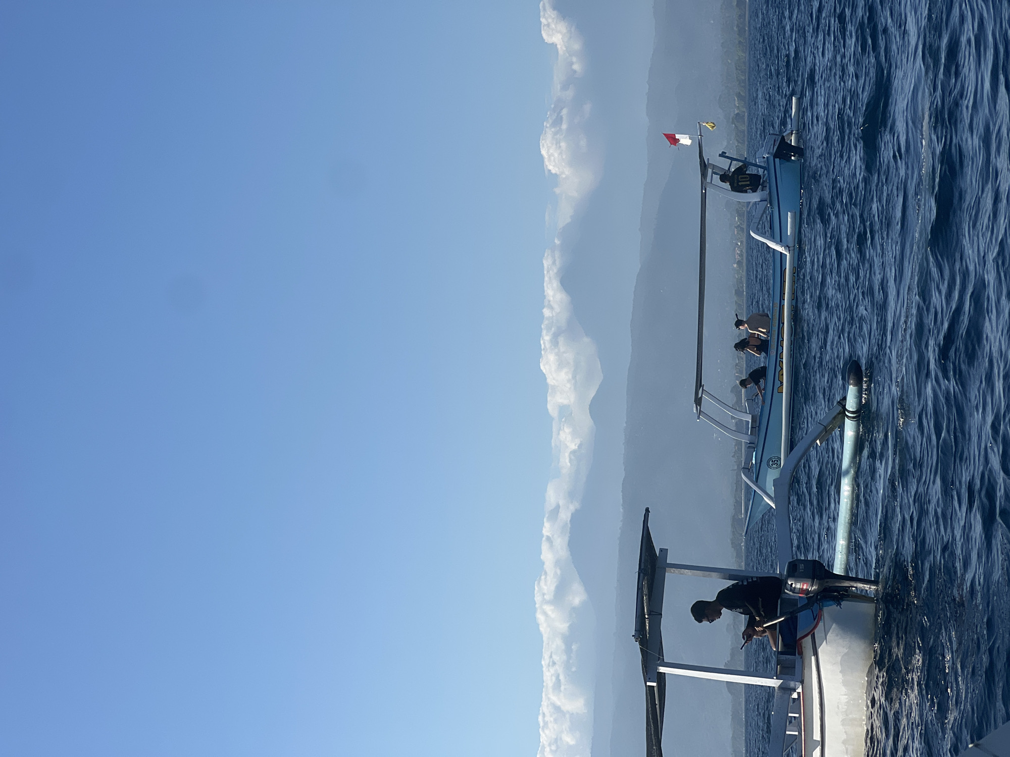 The image depicts a serene ocean scene with two traditional Indonesian fishing boats in the foreground, situated in the waters off the coast of Seririt, Indonesia. The sky is a clear, bright blue, with a few wispy clouds forming a band above distant, hazy mountains. The water is a deep blue, with visible ripples indicating a gentle breeze.
On the left, a white and blue fishing boat has a covered canopy. A man wearing sunglasses and a black long-sleeved shirt sits on the edge of the boat, looking down at his phone. An outboard motor is visible on the back of the boat.
To the right, a lighter blue fishing boat carries several passengers. One person in a black shirt with the number "10" on the back stands at the stern, appearing to steer. A red and white Indonesian flag flutters from a pole at the rear of this boat, and a small yellow fish ornament hangs from the same pole. Several people are seated under the canopy of this boat.
The background reveals a range of mountains, some silhouetted against the sky, others partially obscured by clouds. The overall impression is one of a calm, bright day at sea, likely for leisure or transport.