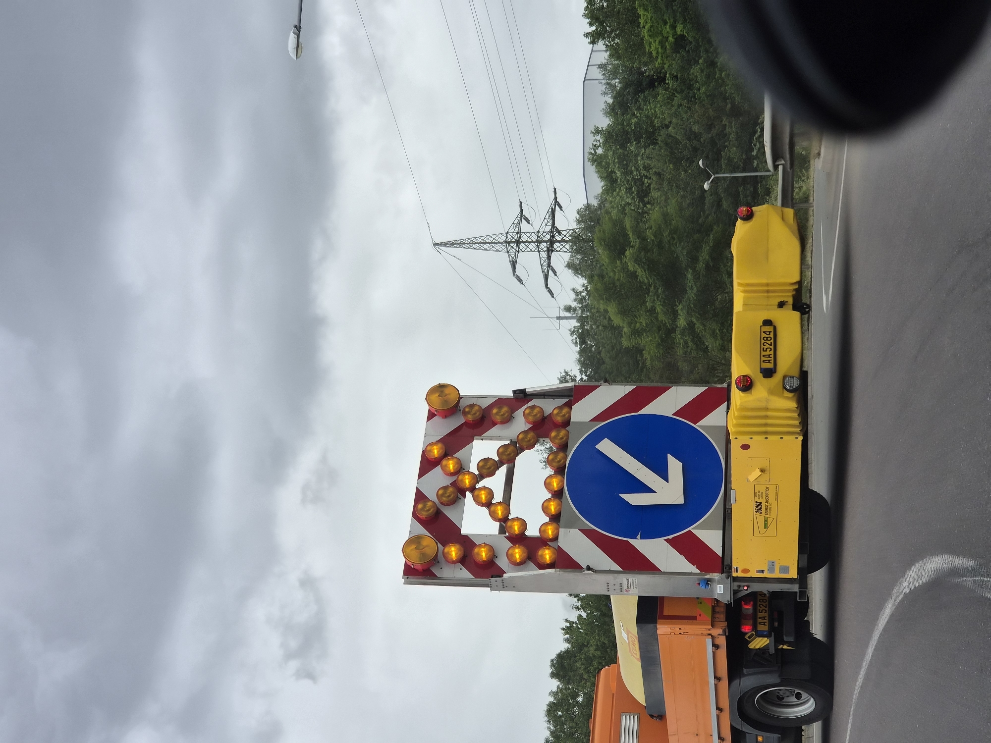 The image displays the rear section of a road maintenance vehicle on an asphalt road in Sanem, Luxembourg, under an overcast sky. The vehicle is an orange truck equipped with a large, rectangular warning sign and a yellow impact attenuator.
The warning sign features alternating red and white diagonal stripes. Its upper portion contains numerous amber circular lights arranged in a chevron configuration pointing right, with additional amber lights along the perimeter. Below this, a circular blue sign with a thick white arrow pointing diagonally down and to the left is displayed. The brand name "horizont" is visible on the sign's support structure. An orange truck bed is partially visible, containing a yellow tank labeled "CEMO." The vehicle's license plate, "AA 5284," is present on the truck's rear bumper and also on the yellow impact attenuator. The impact attenuator, a crash cushion device, is affixed to the vehicle's rearmost section, displaying the text "3500X NCHRP 350 TL-3 ENERGY ABSORPTION SYSTEMS, INC." and integrated rear taillights.
The road surface exhibits white curved markings. A metallic guardrail is partially visible on the right side of the road. Dense green trees and foliage line the background. Above the tree line, a large metallic electricity transmission pylon stands prominently, supporting multiple overhead power lines. A large, light-colored industrial building is partially visible behind the trees to the right of the pylon. Two streetlights are also present: one in the mid-ground on the far right and another further back within the treeline. The sky is characterized by an expanse of grey clouds.