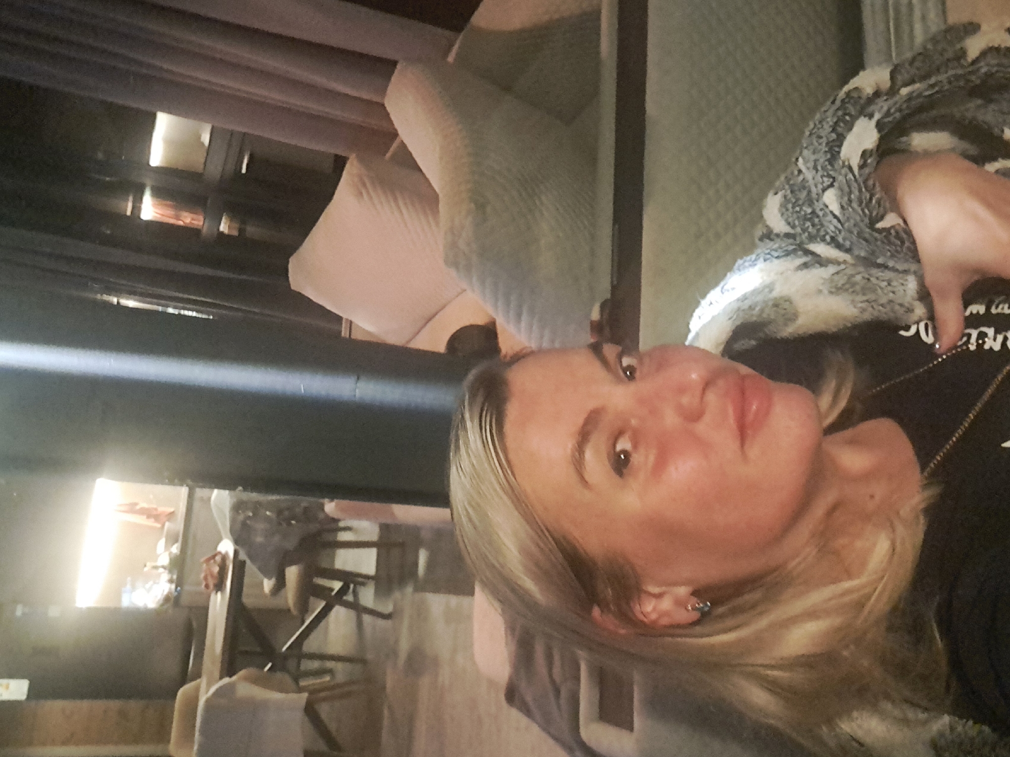 The image shows a woman with blonde hair, wearing a black t-shirt and a patterned scarf, leaning towards the camera. She is seated in what appears to be a living area with furniture including a couch with plush pillows and a dining area with a table and chairs visible in the background. The lighting suggests an indoor evening setting. The location of Krasnyi Ogorok, Russia is not discernible from the visual information provided in the image.