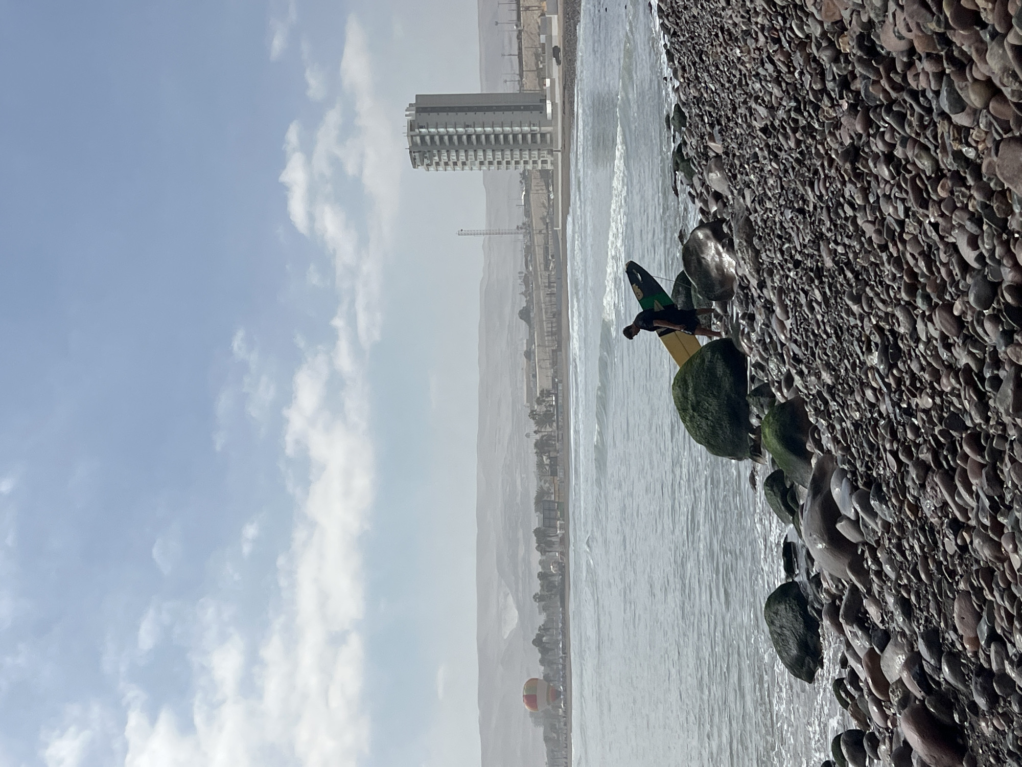 This image shows a rocky beach on the coast of Arica, Chile. The beach is composed of small, smooth stones of varying colors. A man stands on the rocks near the water's edge, holding a surfboard with green and black accents. The water is turbulent with visible waves. In the background, a cityscape is partially obscured by haze, featuring a tall, multi-story building and a construction crane. The sky is overcast with scattered clouds.