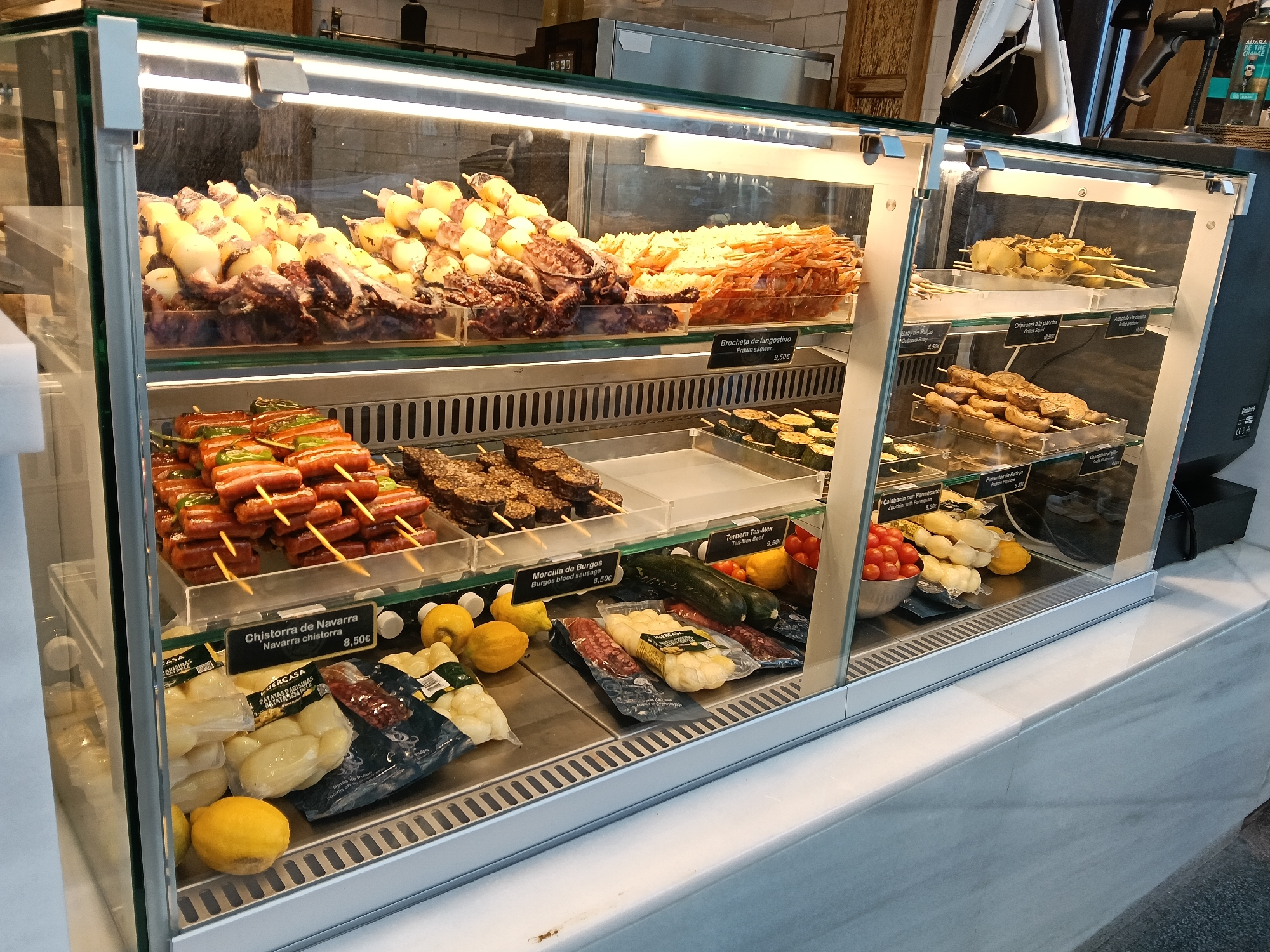 A refrigerated display case presents an assortment of food items in a retail setting, potentially a food market or deli, located in Madrid, Spain. The case is filled with various skewered meats and seafood, including what appears to be octopus, prawns, squid, and a red meat sausage known as chistorra. Other items in the case include packages of potatoes, zucchini, and various skewers of meat and vegetables. Price tags are visible for most items. The case is well-lit, emphasizing the food's presentation. The counter beneath the case is made of marble.