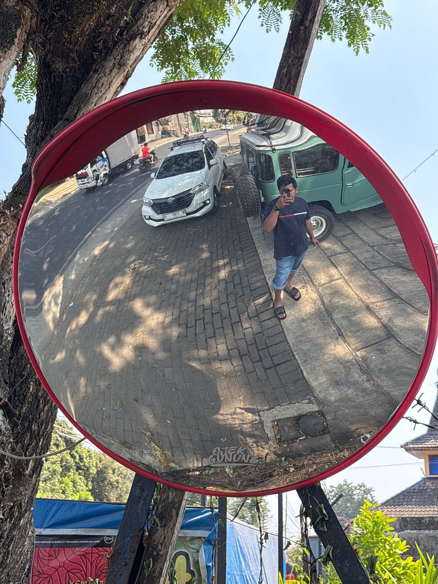 In Belung, Indonesia, a convex traffic mirror, framed in red, reflects a sun-drenched street scene. A man in a navy blue t-shirt and denim shorts stands in the reflection, holding a smartphone and seemingly taking a selfie. To his left, a white SUV is parked on a brick-paved road, while a vintage green truck sits behind him. The mirror is mounted on a metal pole, attached to a tree with thick, textured bark. The background shows a clear blue sky, lush green foliage, and parts of buildings.