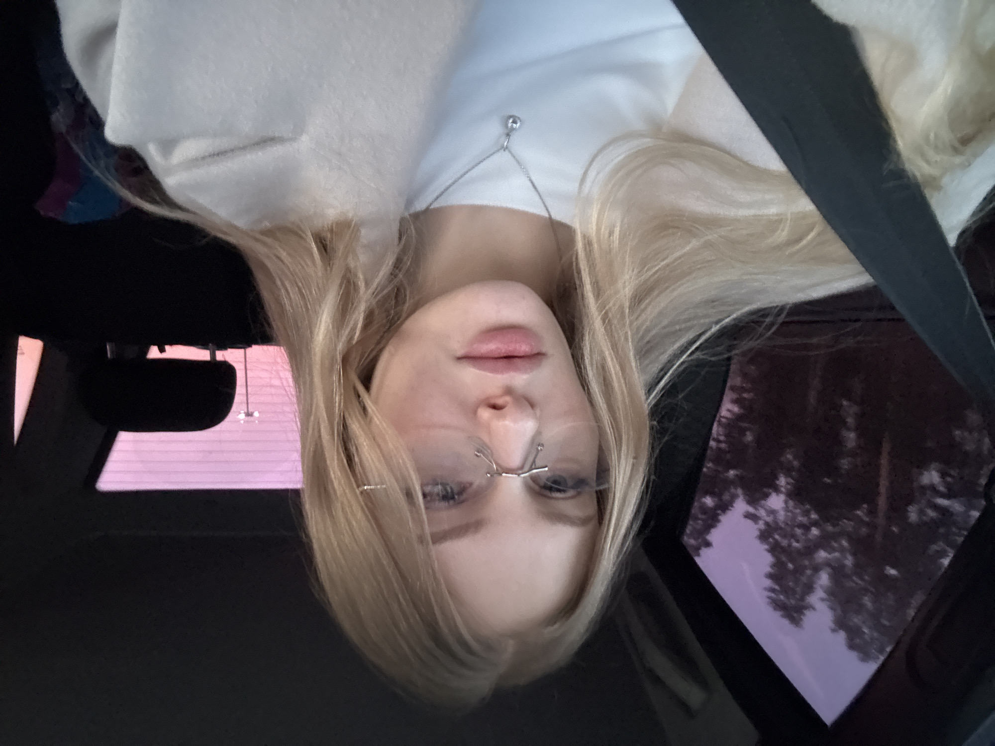 A woman with blonde hair and glasses is in a car, wearing a white shirt and a light-colored jacket. A seatbelt crosses her chest. The car's sunroof shows a view of trees and a purple sky. This image was taken in the city of Perm, Russia.