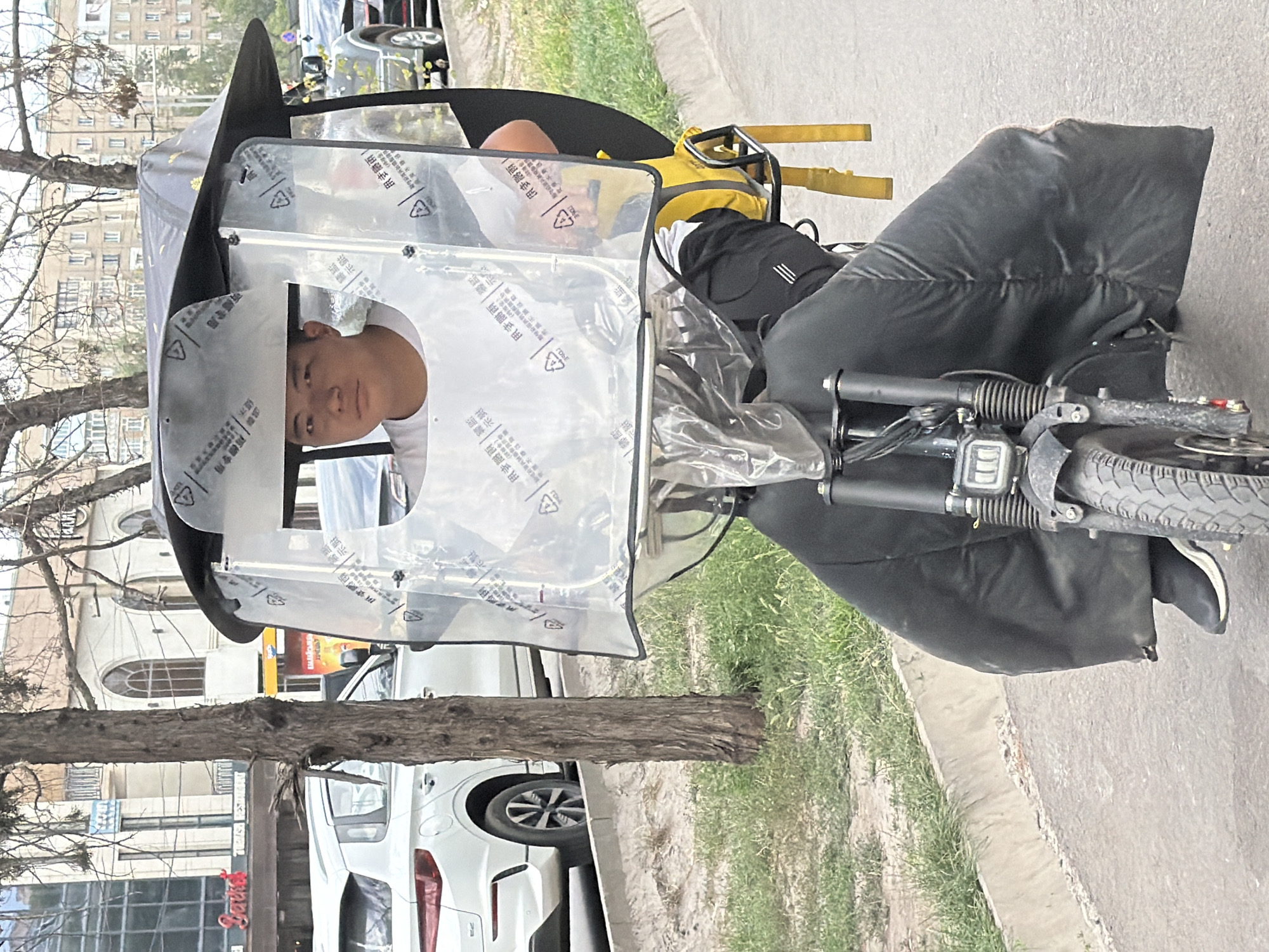 A young East Asian man with short dark hair and a neutral expression is riding an electric scooter on a paved street in an urban environment. He is wearing a white t-shirt and dark pants. The scooter has a clear plastic enclosure with a black frame around it, providing some protection from the elements. The enclosure is covered in Chinese characters. The man is wearing a black padded cover that extends from his waist down, obscuring his legs and the lower part of the scooter. He has a yellow insulated delivery bag attached to the back of the scooter. In the background, there are trees, buildings with arched windows, and other vehicles. The lighting suggests it is daytime, possibly overcast.