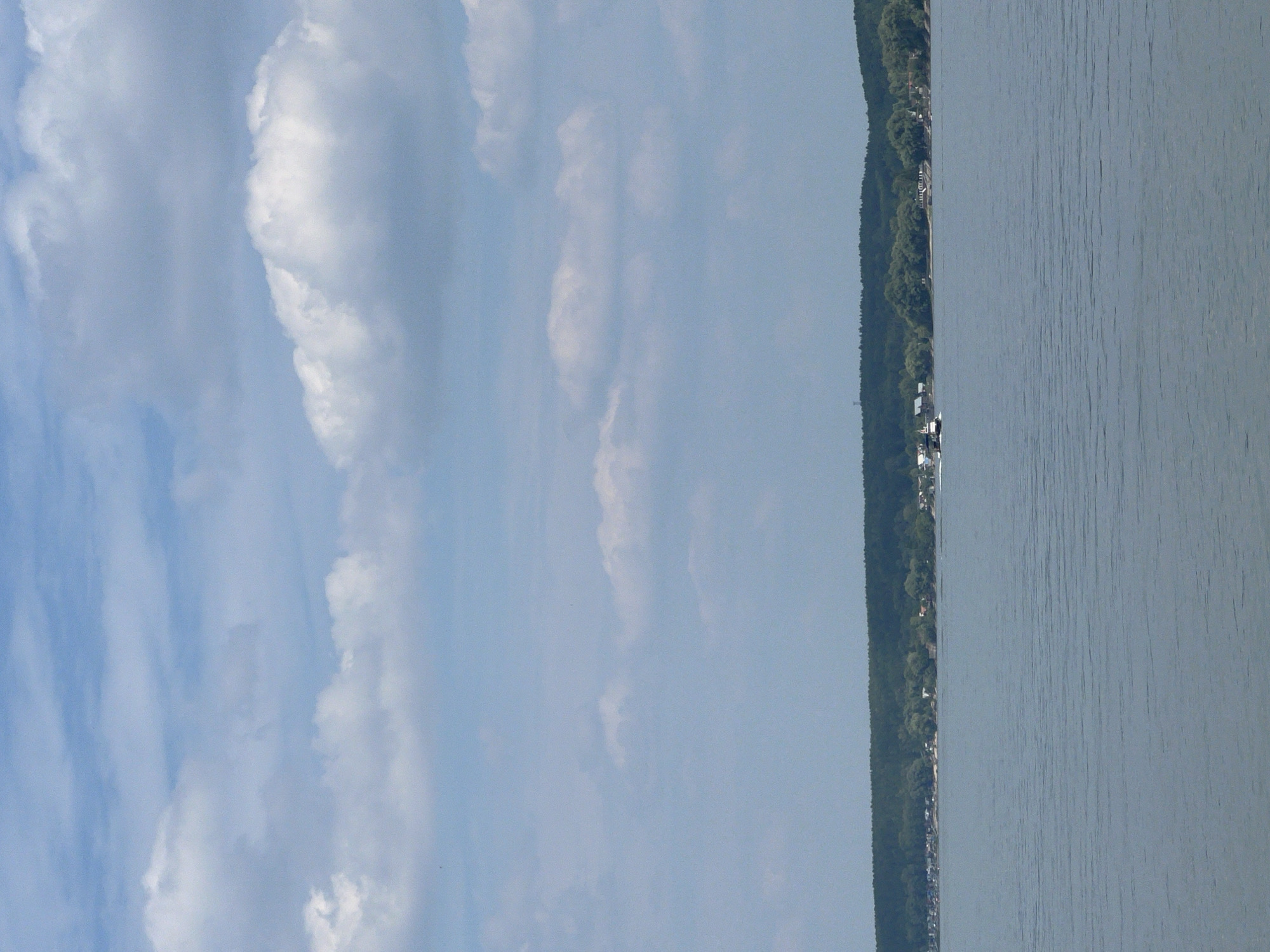 The image shows a wide body of water, likely a river or lake, under a partially cloudy sky. The water has ripples, indicating some movement. Along the far bank of the water, there is a dense line of trees, and beyond that, scattered buildings can be seen, suggesting a settlement. A small boat is visible in the distance on the water, moving towards the left. The sky is a pale blue with large, fluffy white clouds. The overall impression is a serene, natural landscape with signs of human habitation in the distance. This scene is consistent with the location of Mochishche, Russia, which is situated along a body of water and features forested areas and settlements.