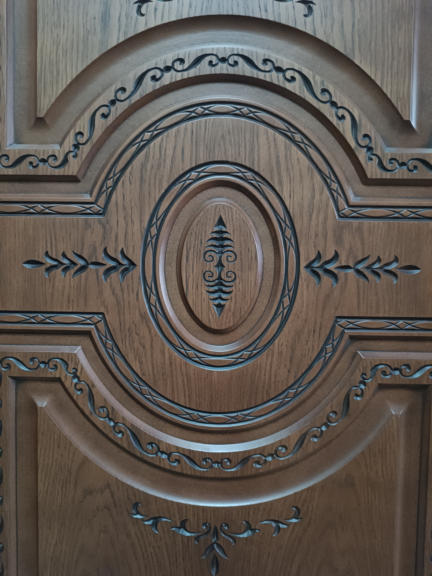 The image shows a close-up view of a carved wooden door. The door features an ornate central oval panel with intricate scrolling designs and a leaf-like motif in the center. Surrounding the central panel are concentric carved circles and rectangular sections with decorative borders and stylized leaf patterns. The wood grain is visible, and the carving is dark, creating a contrast with the lighter wood. The image appears to be a detail shot of a decorative element on a door, likely an entrance door to a building or a significant internal door. There are no people or environmental context visible in the image, other than the implied setting of a building interior or exterior where such a door would be found. The location in Myrza-Ake, Kyrgyzstan, is not visually represented in the photograph itself.