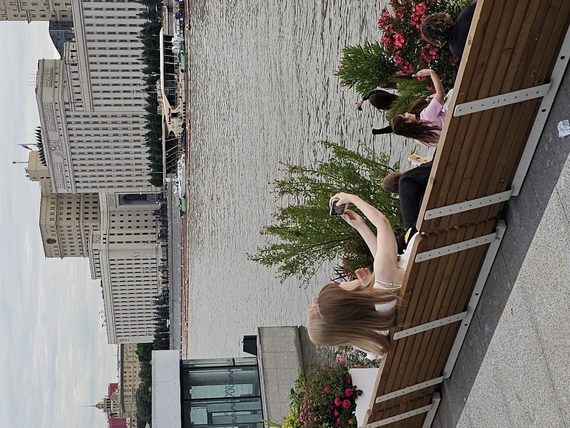 The image depicts a sunny day with scattered clouds in Moscow, Russia. The scene is a waterfront, with a wide river or canal in the background. On the left side of the frame, large, imposing buildings with many windows line the embankment, suggesting an urban environment. One of these buildings has a Russian flag flying.
In the foreground, a group of people are seated on a long, wooden bench that is positioned next to the water. The bench has a modern design with white metal supports. Several individuals are visible, including a young woman in a light pink top and a person in a black top with curly hair. Another young woman in a white top is taking a selfie with her phone. The people appear to be casually enjoying the outdoor setting.
Behind the bench, there are lush green plants and vibrant pink flowers, adding a touch of nature to the urban landscape. The water reflects the sky and buildings, creating a sense of depth and tranquility. The overall atmosphere seems relaxed and pleasant, indicative of a leisurely day by the water. No visible text is present in the image.