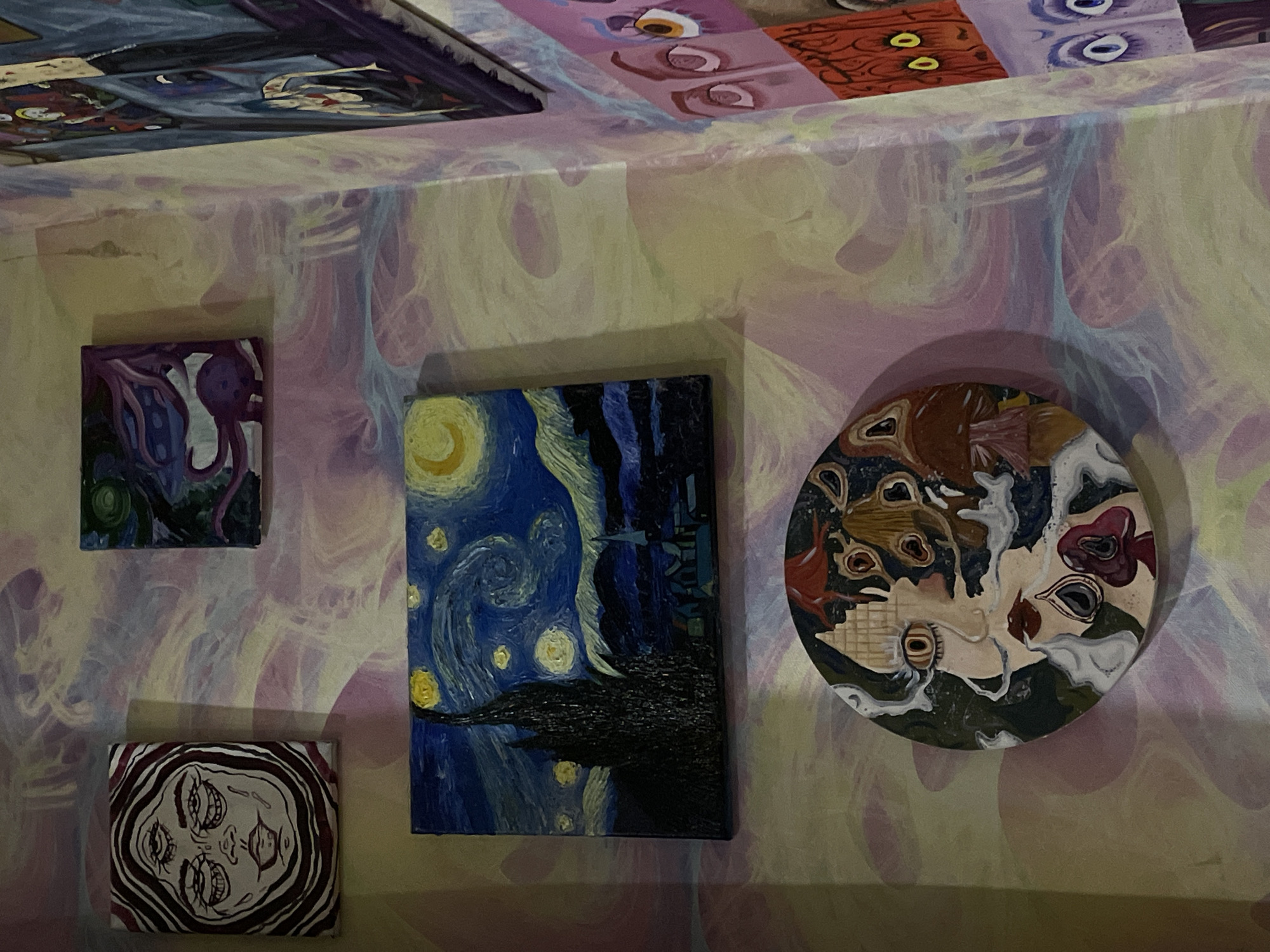 The image shows a wall in an apartment decorated with several paintings. The wall has a swirled pattern in pastel colors. On the wall, there are at least five paintings. From top to bottom and left to right:
1. A small rectangular painting depicting a face with three eyes, drawn in black lines with red accents, on a white background with red stripes.
2. A small square painting with a purple creature in a dark, abstract landscape.
3. A rectangular painting that appears to be a rendition of Van Gogh's "The Starry Night".
4. A circular painting with a collage-like image of a face with one eye visible, surrounded by swirling elements and possibly mushroom motifs.
5. A portion of a painting on the right side of the image, which shows several stylized eyes on a pink background.
There is no indication of the location in the city of ЖК «Борисоглебское», Russia. The image is taken indoors, likely in a residential setting. There are no people visible in the image.