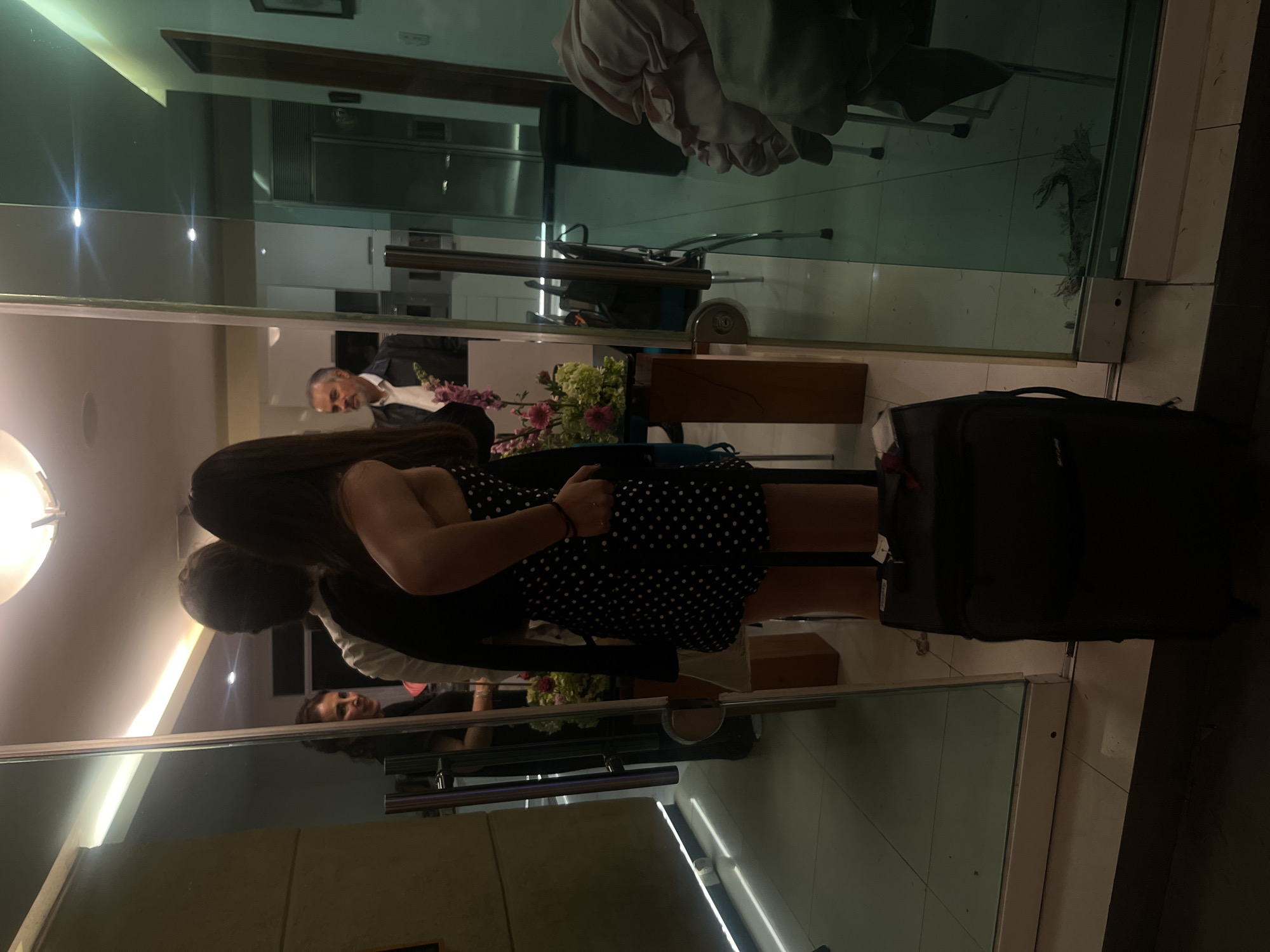 The image depicts a reflection through a glass door. Inside, a man in a dark suit and a white shirt stands behind a table with flowers. He appears to be speaking or greeting someone. A woman in a black and white polka-dot dress stands closer to the door, obscuring much of the interior. A black suitcase rests on the tiled floor near the doorway. The setting appears to be an entryway or lobby of a building. The location is identified as San Bernardino Tlaxcalancingo, Mexico.