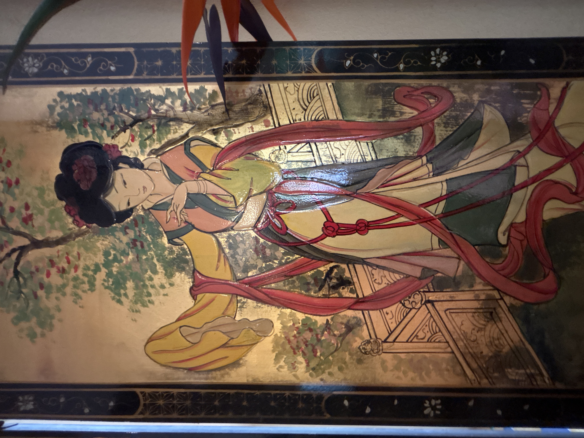 The image depicts a vertical, ornate painting on what appears to be a panel or screen, featuring a traditional East Asian woman in an elaborate, flowing robe. The artwork has a metallic, likely gold, background, with dark blue borders on the left and right sides, decorated with intricate patterns. The woman, with her hair styled in an updo adorned with red flowers, wears a yellow and pink garment with green accents, draped with long, flowing red sashes. She is depicted with delicate facial features and her hands are posed gracefully. Behind her, a stylized tree with green foliage and pink blossoms suggests a garden setting. In the foreground, a decorative railing or fence is visible. The overall style is painterly with bold outlines and vibrant colors.
The location provided, "Chalong, Thailand," is not visually represented within the image itself. The painting is the sole subject, and there is no contextual information within the frame to suggest a specific city or geographical location. The painting's style is characteristic of East Asian art, not Thai art, further decoupling it from the specified location.