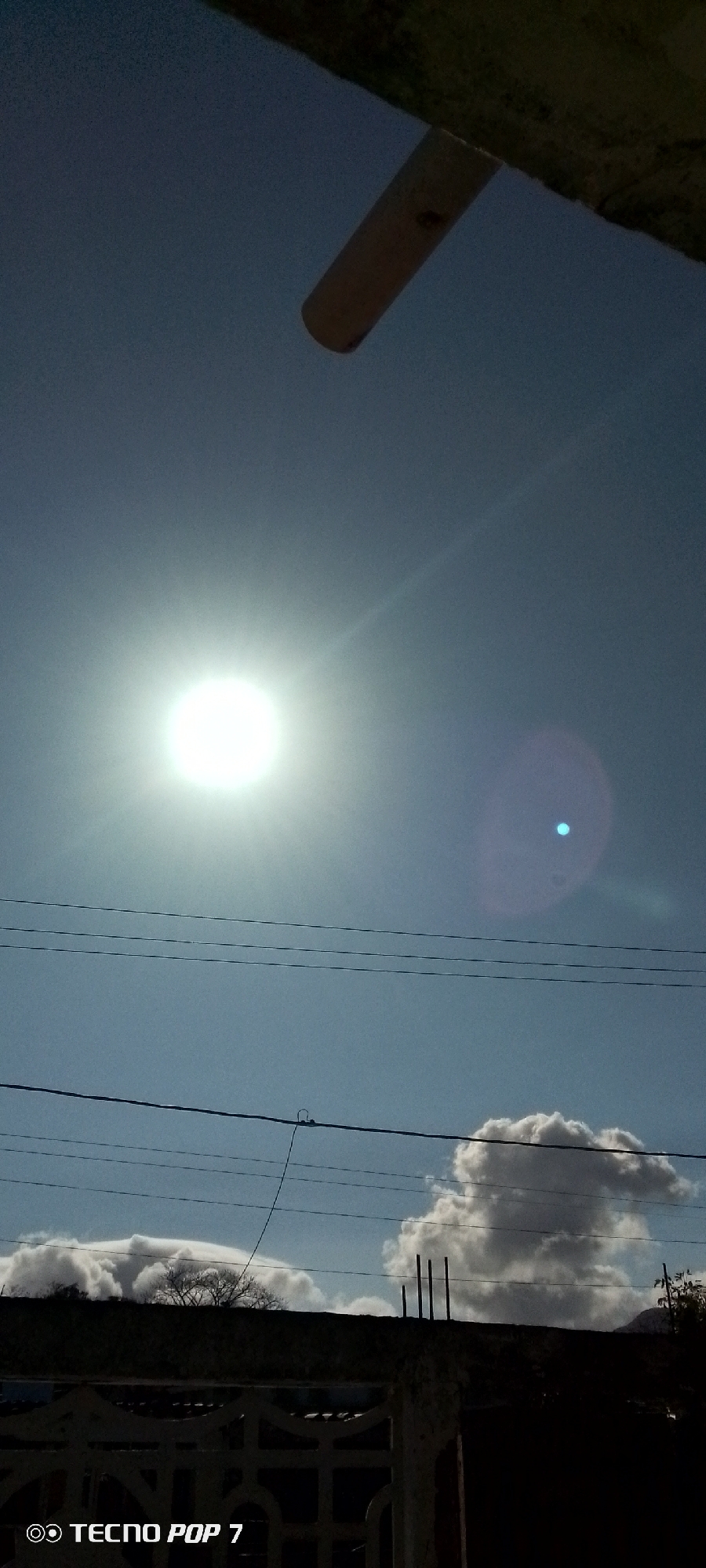 The image captures a bright, sunny outdoor scene under a clear blue sky. A brilliant sun is prominently featured in the upper left, casting an intense glare and producing a distinct blue lens flare towards the upper right. Multiple dark power lines stretch horizontally across the middle of the frame. Below these lines, white cumulus clouds are visible along the horizon, forming distinct puffy formations, particularly on the right side.
The lower portion of the frame is dominated by the dark silhouette of a low-lying structure, likely a wall or fence, from which several rebar rods extend upwards on the right. Sparse dark vegetation is visible above this structure, mainly on the left. An unseen, dark structure partially covers the top of the frame, from which a light-colored cylindrical object hangs downwards.
The photo appears to be taken around midday, indicated by the high position and intensity of the sun. The weather is clear and bright. In the bottom left corner, a watermark reads "◎ TECNO POP 7." The setting suggests a residential or semi-urban environment, potentially in Altagracia, Venezuela, given the context. No specific activity or people are visible; the scene focuses on the interplay of natural light and man-made structures.