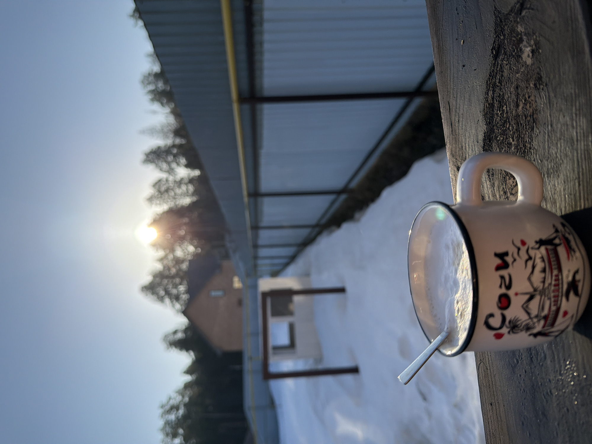 The image shows a mug with coffee on a wooden post in Chaikovsky, Russia. The mug is white with a "COZY" design. There is a spoon inside the mug. The background shows a snowy landscape and a building. The sun is shining brightly. The image is taken outside.