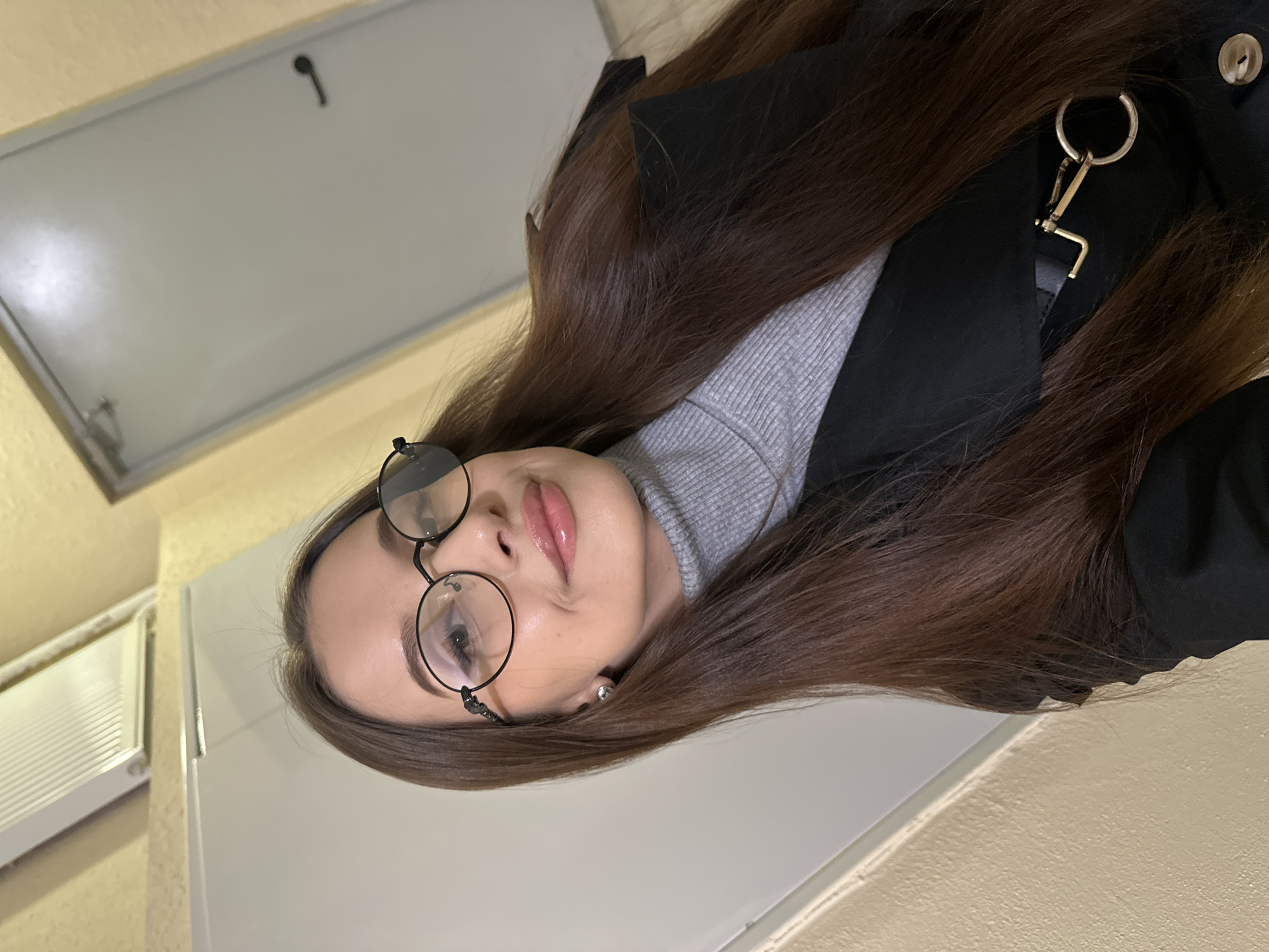A young woman with long, brown hair, wearing round glasses, a grey turtleneck sweater, and a black coat, is positioned in front of a plain white wall. A metal object hangs from her coat. The environment includes a white air conditioning unit on a yellow wall in the background, and a metal door handle on a white panel in the upper left corner. There is no information available to establish the specific location in Yekaterinburg, Russia.