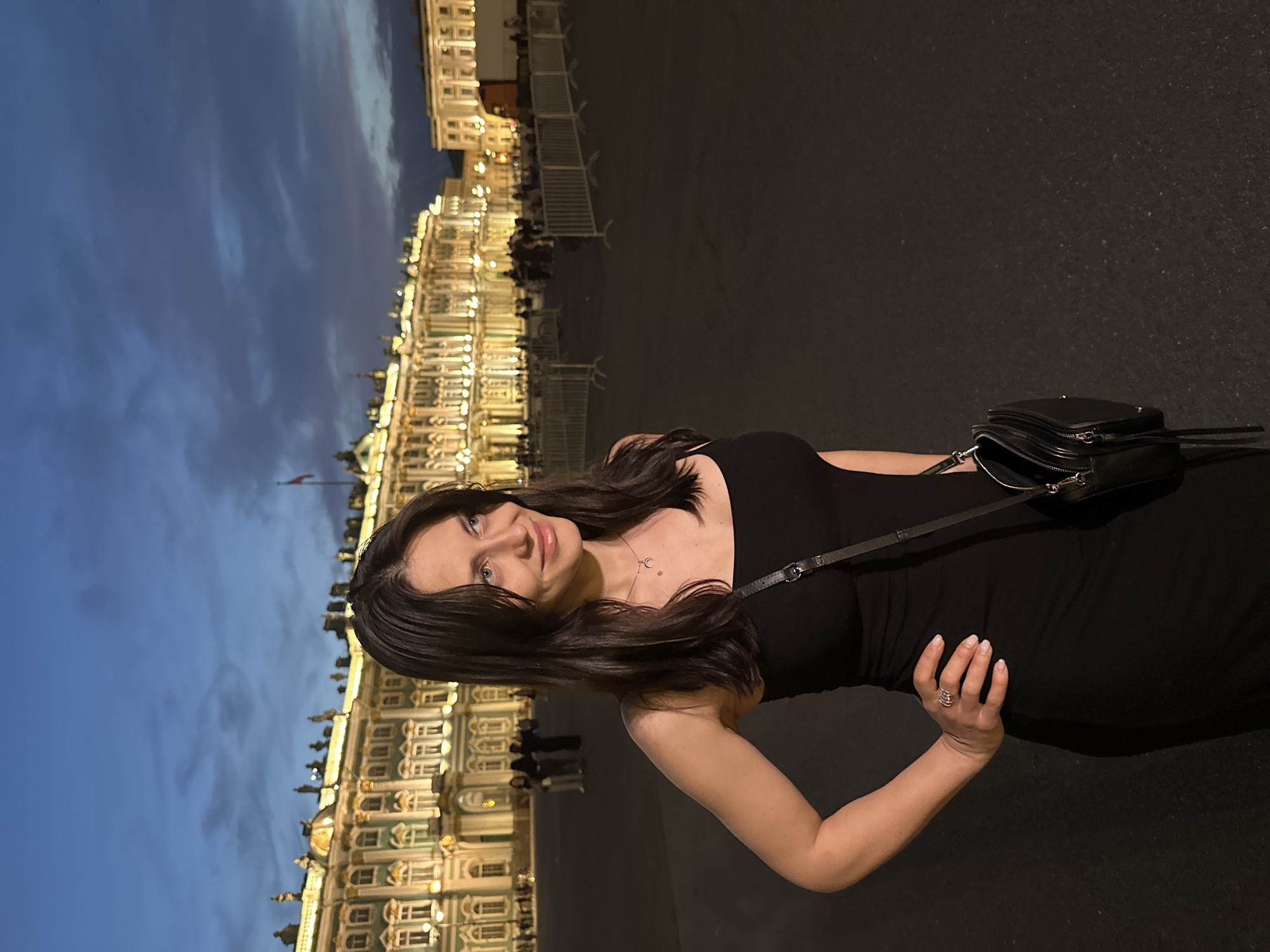 A young woman with long, dark hair and blue eyes, wearing a black sleeveless dress and a silver necklace, stands in a public square at dusk. She has one hand on her hip and a black crossbody bag is slung over her shoulder. In the background, the Palace Square in Saint Petersburg, Russia, is illuminated by artificial lights, showcasing the grandeur of the Winter Palace and the General Staff Building. The sky is a deep blue with hints of twilight. Several people and a truck are visible in the distance.