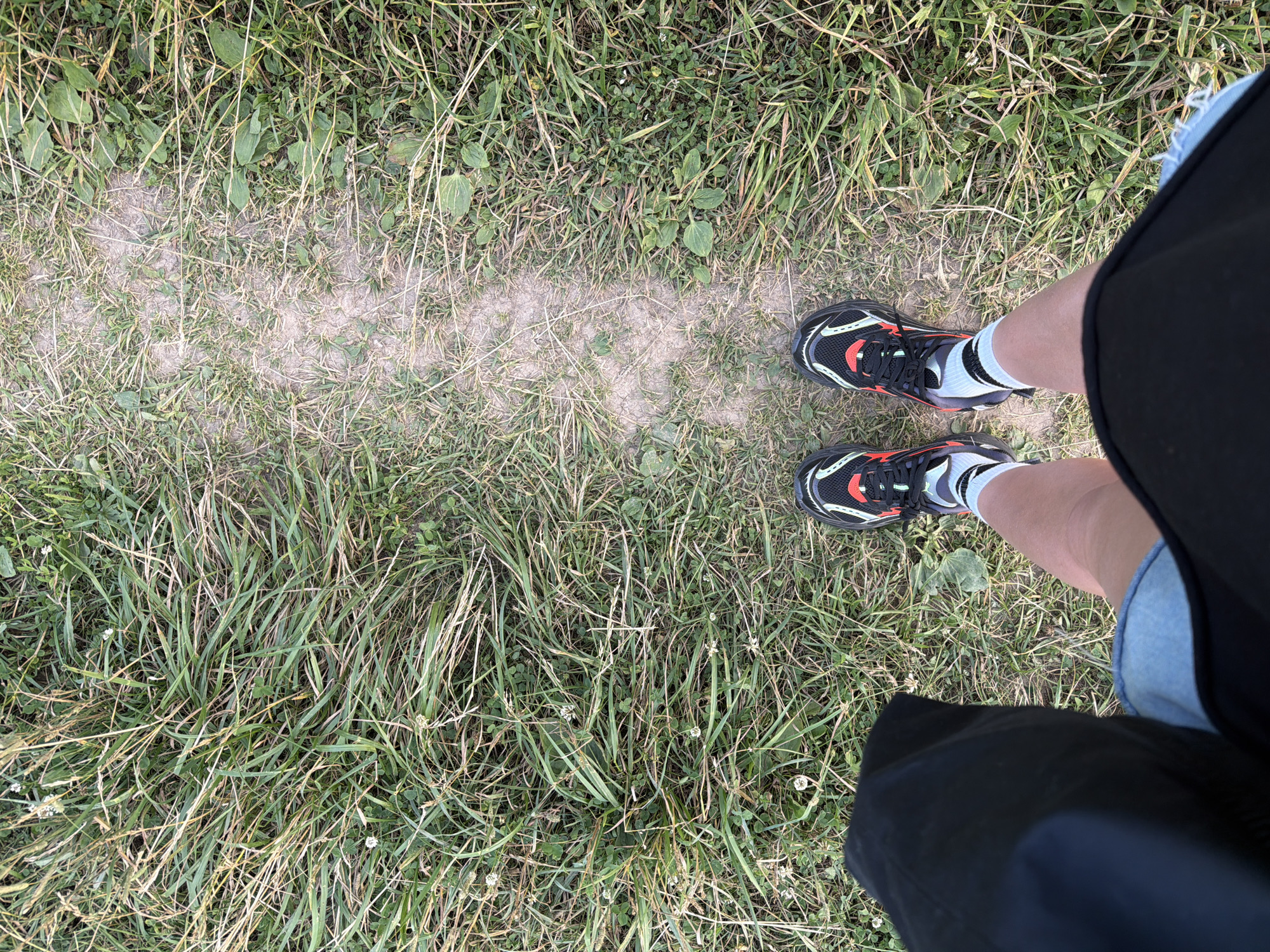 A first-person perspective shot captures a person standing on a dirt path surrounded by grass. The person is wearing black athletic shoes with red and white accents, and light-colored socks with black stripes. Denim shorts and a black skirt or shorts are visible. The scene appears to be outdoors in a natural setting, possibly a park or trail, with the sun casting a light shadow on the path, suggesting it is daytime. The grass is green and somewhat overgrown, with scattered small white flowers. The ground is a mix of dirt and grass. The overall impression is casual and active, likely taken during a walk or outdoor activity. No text is visible in the image. The location context provided is Mar'inskoe, Russia.