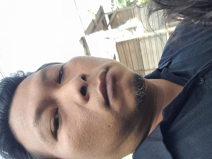 A man in a black collared shirt is close to the camera, his face angled toward the viewer. He has short dark hair and a sparse, graying beard. The background is blurred, showing a building with corrugated metal walls and glimpses of greenery. The image appears to be taken in daylight, possibly outdoors, with the camera angled upward toward the subject.