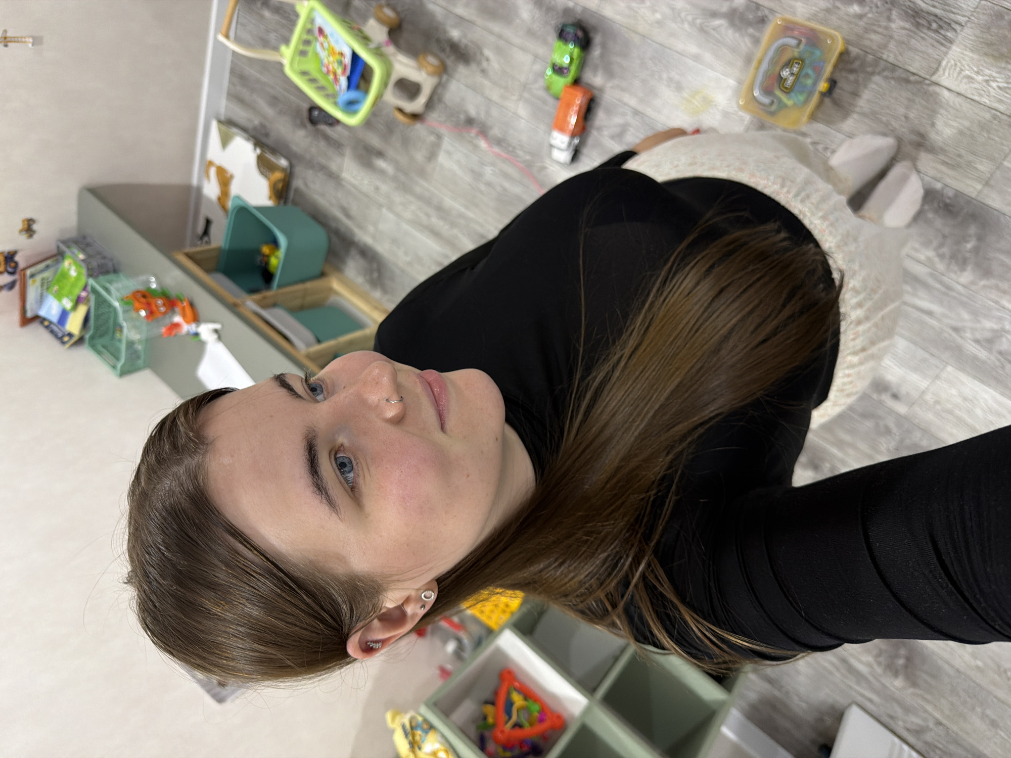 A young woman with long, brown hair is captured in a high-angle, indoor shot. She is wearing a black turtleneck top and white patterned leggings. Her facial features are visible, with blue eyes, a nose ring, and piercings on her left ear. The background suggests a playroom or child-friendly area, with various toys scattered around, including a small green shopping cart, toy cars, a shelf unit filled with toys, and a storage bench with containers. The floor appears to be made of grey laminate or wood-effect tiles. The overall environment suggests a residential setting, likely a home in Chaikovsky, Russia, designed for children's activities.