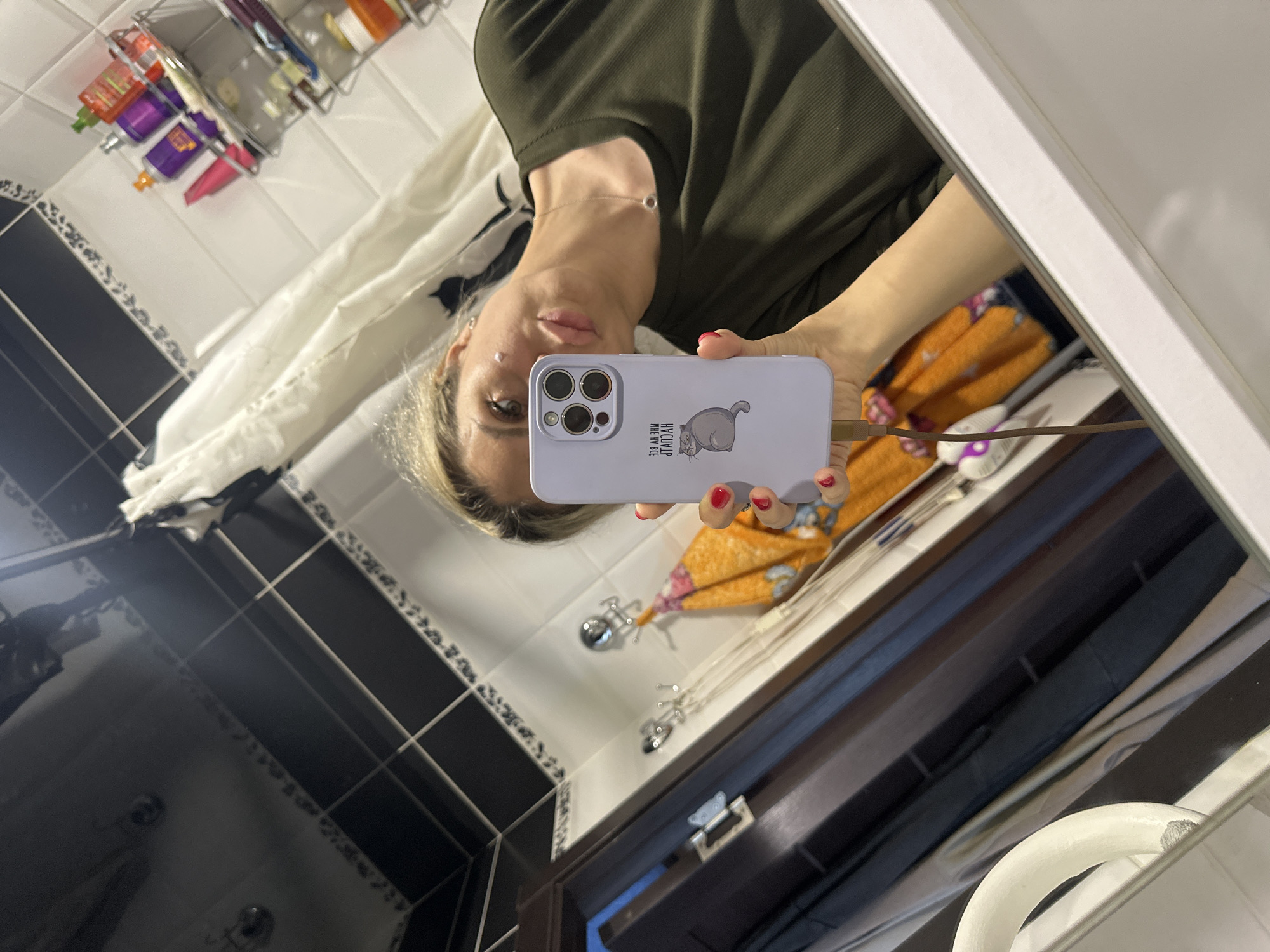 The image is a mirror selfie taken in a bathroom. A woman with blonde hair is holding a light purple smartphone in her left hand, which is reflecting in the mirror. She is wearing a dark green long-sleeved shirt and a thin silver necklace with a circular pendant. The bathroom has white tiled walls with black grout lines, and a black tiled ceiling that reflects the surroundings. On a shelf in the background, there are several bottles of toiletries, including a red, a purple, and an orange bottle. A white shower curtain is visible. To the left of the woman, a dark brown wooden door frame is present, and an orange towel with a floral pattern hangs on hooks. The overall lighting suggests an indoor environment. There is no information to determine the location in Novoaltaysk, Russia.