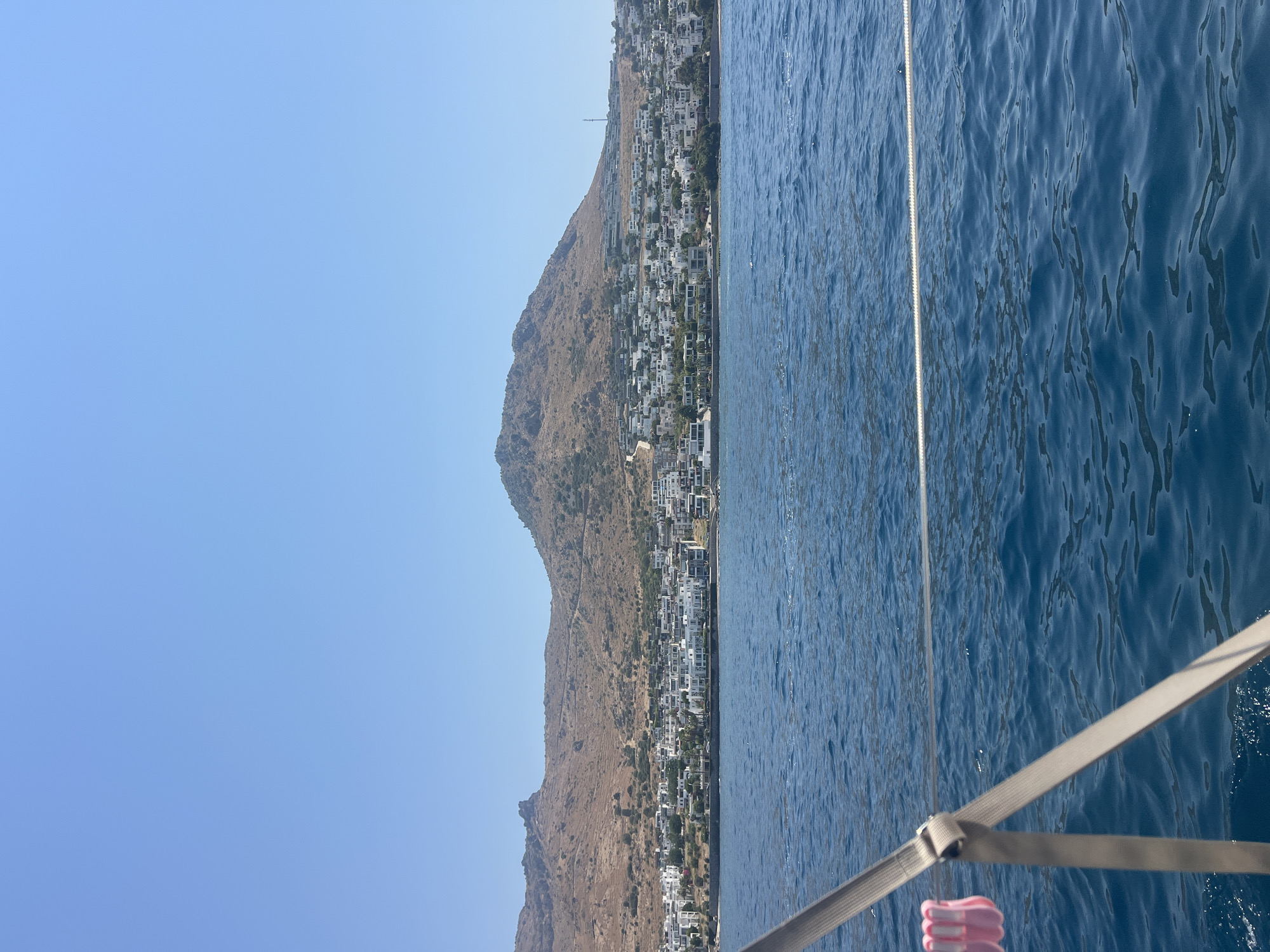 A panoramic view from a boat shows a coastal town nestled at the base of a mountain under a clear blue sky. The town is characterized by numerous white buildings with red-tiled roofs, densely packed along the shoreline and climbing up the lower slopes of the mountain. The mountain itself is arid and sparsely vegetated, with dry, yellowish-brown terrain. The foreground is dominated by the deep blue, rippling water of the sea, with a portion of a boat's railing and a rope visible on the left side, extending diagonally across the frame. Pink clips are attached to the rope. The scene depicts a sunny day with good visibility, suggesting a warm climate.