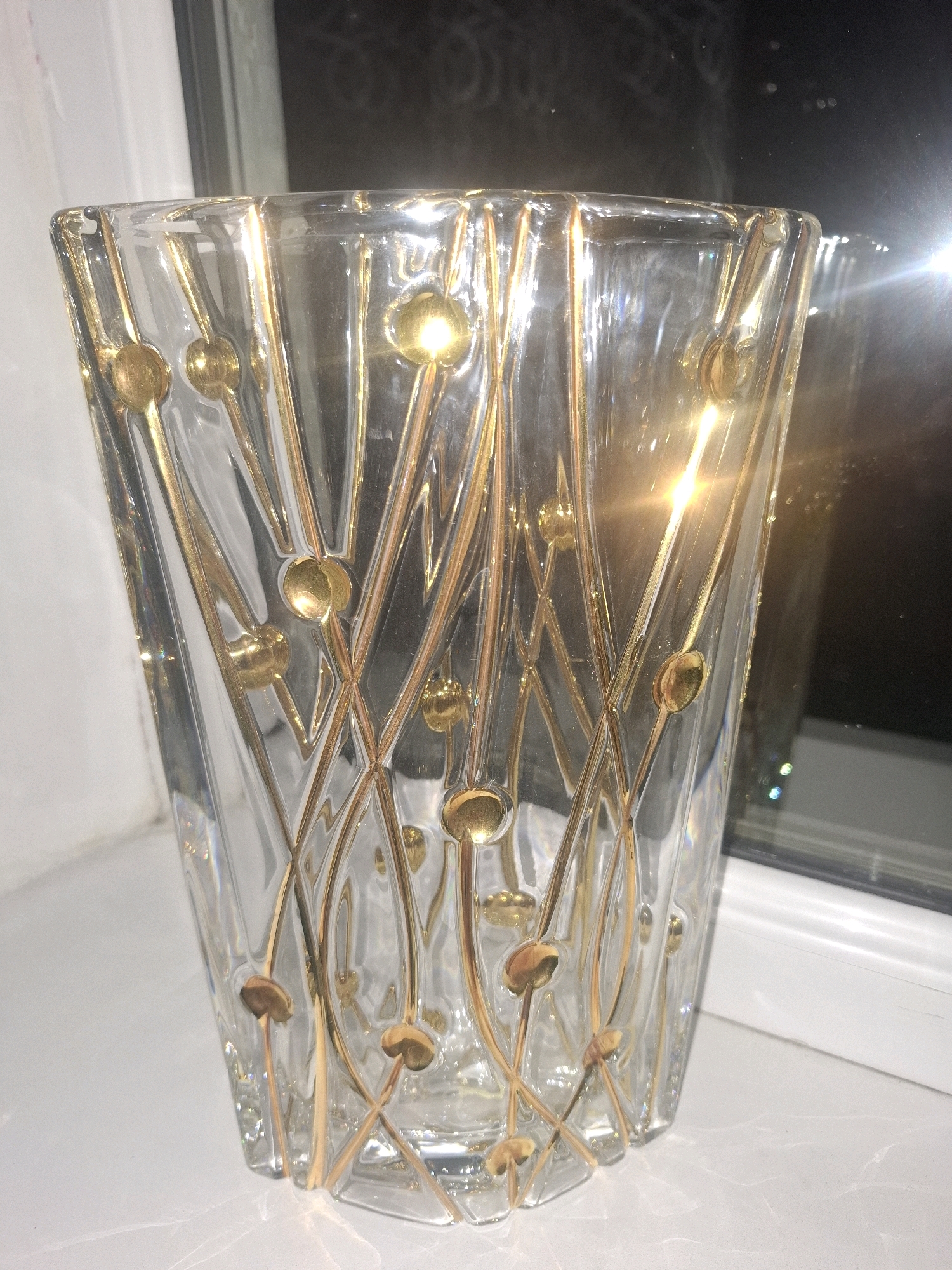 A clear glass vase with gold-colored, abstract decorative lines and dots is positioned on a white, reflective surface. The vase has a rectangular, slightly tapered shape. Light reflects intensely from the top and side of the vase, obscuring some of the details. The background is dark and blurry, suggesting an indoor setting near a window with some light filtering through. There are no people, animals, or specific landmarks visible in the image. The location in Almaty, Kazakhstan cannot be determined from the provided image.