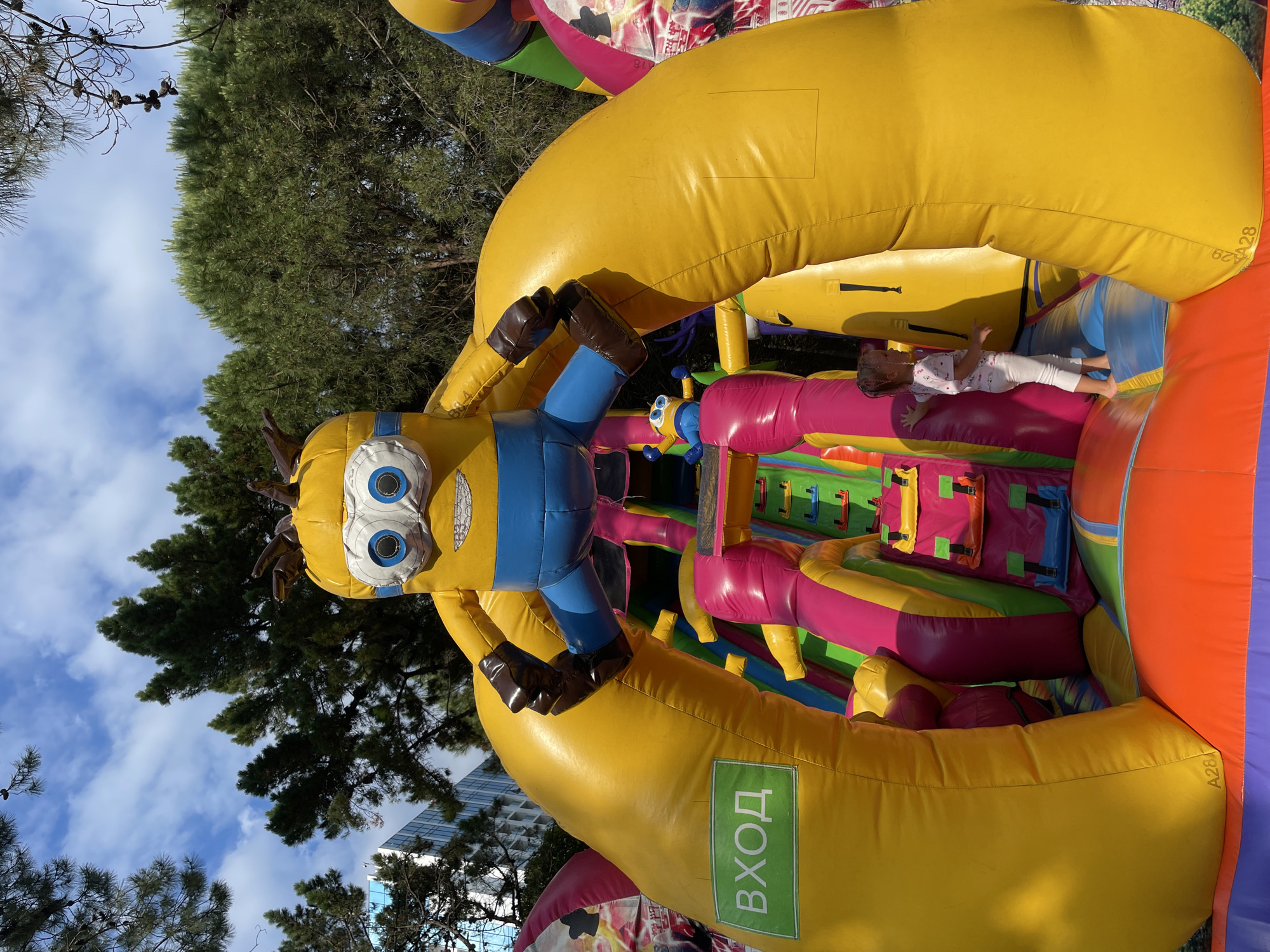 In the city of Sochi, Russia, an outdoor inflatable attraction, resembling a Minion character from the Despicable Me franchise, stands prominently. The large, yellow, inflated structure features the Minion's characteristic blue overalls and goggles, with its arms extended outwards. A young girl, wearing a light-colored t-shirt and pants, is visible inside the inflatable structure, standing on a colorful play area. The background of the image shows lush green trees and a partly cloudy sky, indicating a sunny day. The inflatable structure has a green sign with white Cyrillic text that reads "ВХОД," which translates to "Entrance." The scene appears to be a recreational area, likely a children's play zone.