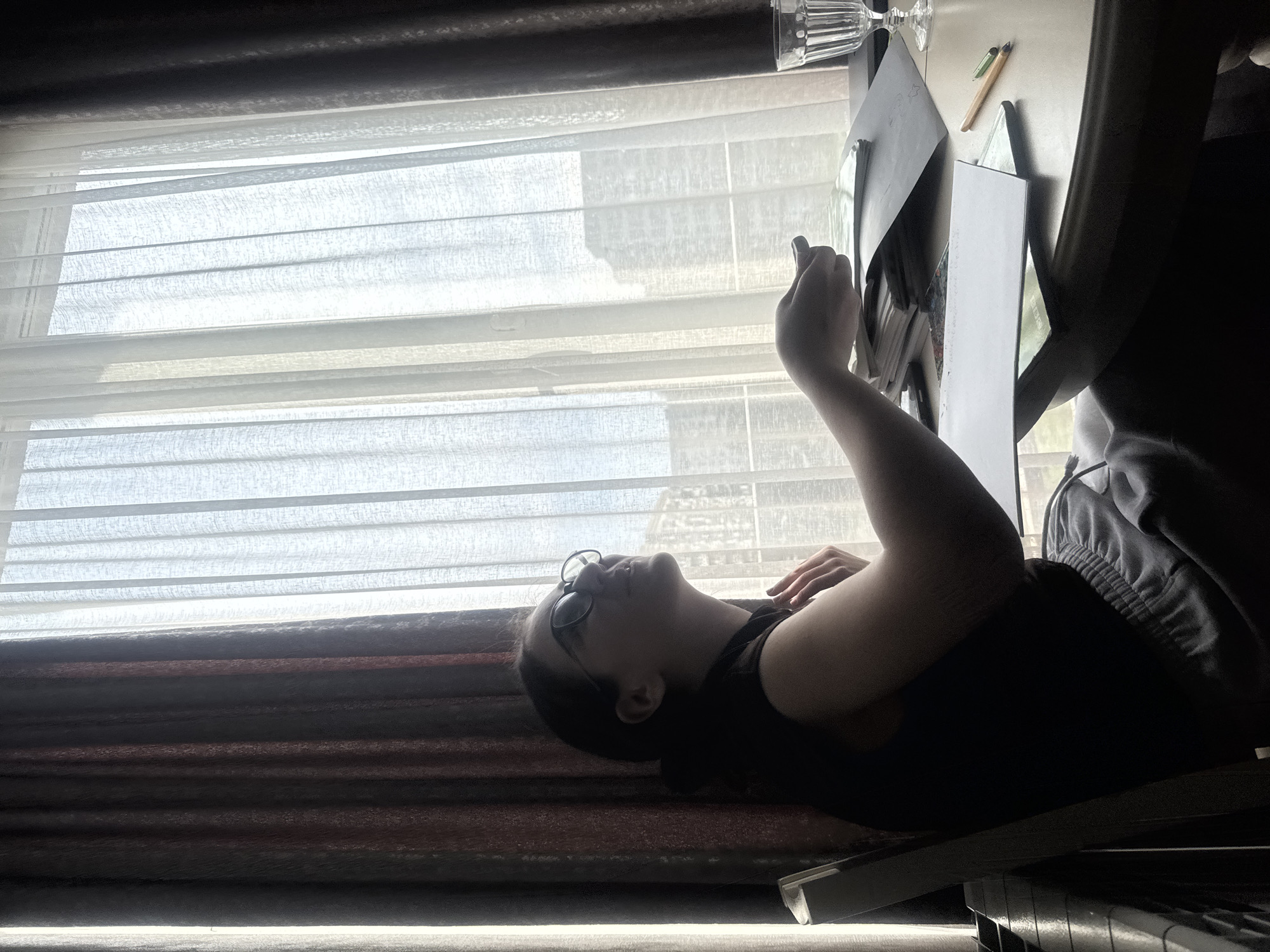 The image depicts an indoor scene in Tbilisi, Georgia, with a person reclining next to a brightly lit window. The perspective is from slightly above, looking down. The original image is rotated 90 degrees counter-clockwise; when oriented correctly, a large window dominates the left side of the frame.
A young person is lying on their back, propped up slightly, facing upwards towards the ceiling. They wear dark-rimmed glasses, a dark short-sleeved top, and grey sweatpants. Their head is positioned closer to the window. The person is actively engaging with a small device, likely a phone or tablet, which they hold in their right hand. Their left arm is relaxed, resting across their stomach or chest.
Behind the person's head, on a desk or table, several items are visible: a clear, cut-glass goblet, a wooden pen or pencil, and multiple papers or small books. The window is covered with white, translucent fabric blinds, which diffuse the bright daylight entering the room. Through the blinds, faint shapes suggest buildings in an urban or suburban setting. Dark, heavy curtains are visible on the far right of the window, framing the bright light. The scene is strongly backlit, casting the person and foreground objects into shadow or silhouette, creating a high-contrast atmosphere typical of daytime with strong window light. There is no visible text in the image. The person appears relaxed, possibly engrossed in their activity.