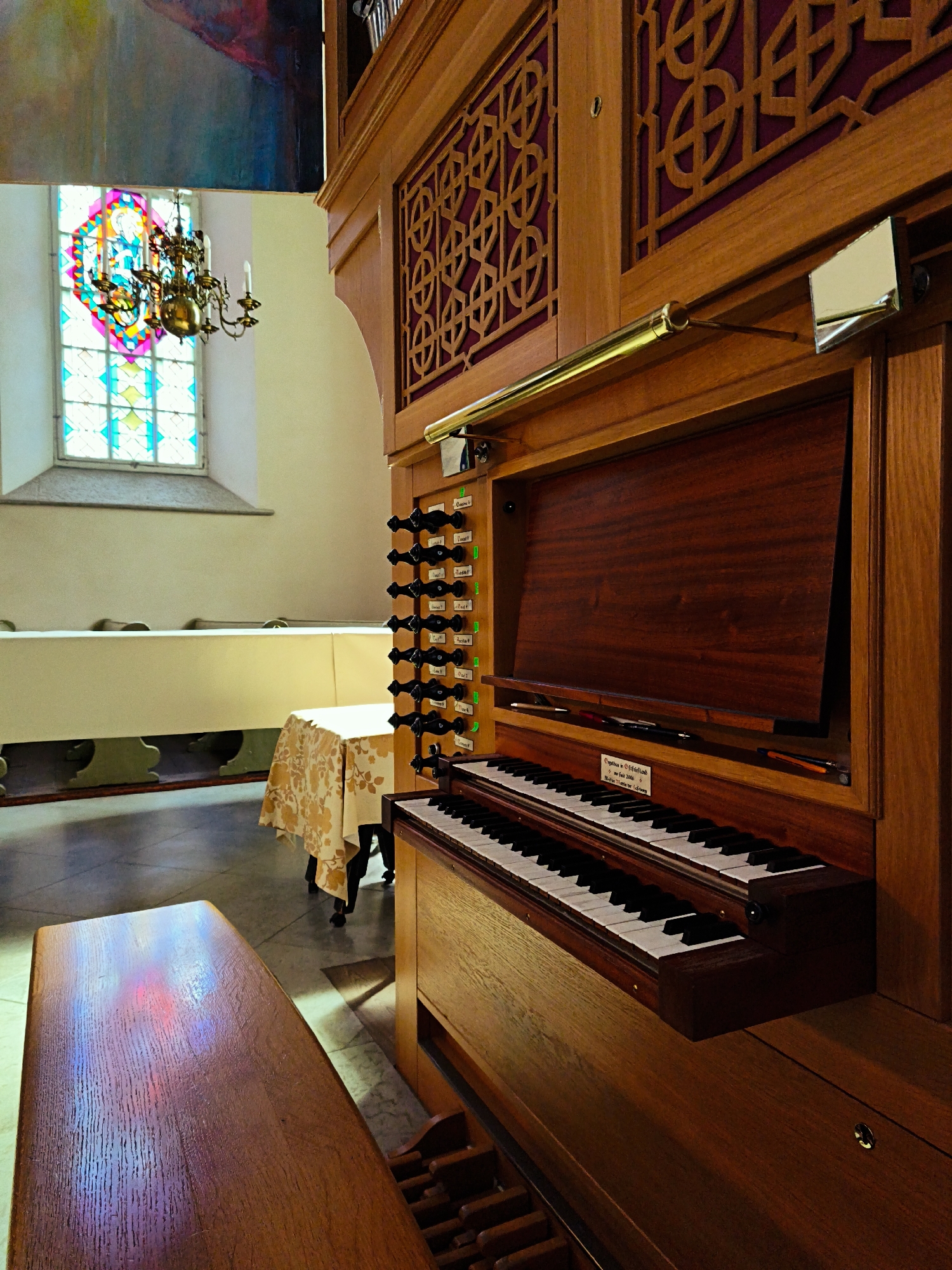 This is an indoor medium shot of a pipe organ in a room that appears to be a church or concert hall in Tallinn, Estonia. The organ, made of polished wood, is the primary subject. In the foreground, the textured wooden bench of the organ is visible, reflecting colorful light from a stained-glass window. The keyboard and stops of the organ are prominent. To the left, a stained-glass window with intricate geometric patterns and bright colors is seen, along with a brass chandelier. In the background, long tables draped in white cloths suggest a dining or gathering space. The overall lighting is warm, with natural light coming through the window illuminating parts of the scene.
