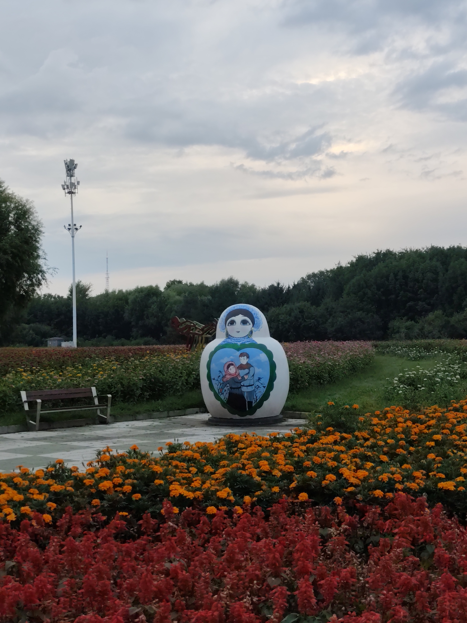 This is a photograph taken in 幸福乡, China, featuring a large, white Matryoshka doll sculpture as the central subject. The sculpture is adorned with a painting of a couple and a child within a heart shape, and the doll itself has a blue headscarf. The doll stands on a paved area in front of a vibrant flower bed. The foreground is dominated by a dense arrangement of red flowers, followed by a layer of bright orange flowers. Behind the doll, a park bench is visible on a checkered paved path. Further in the background, a line of dense green trees forms the horizon, and a tall, white communications tower with multiple antennas is positioned to the left of the doll. The sky above is overcast with soft, diffused light.