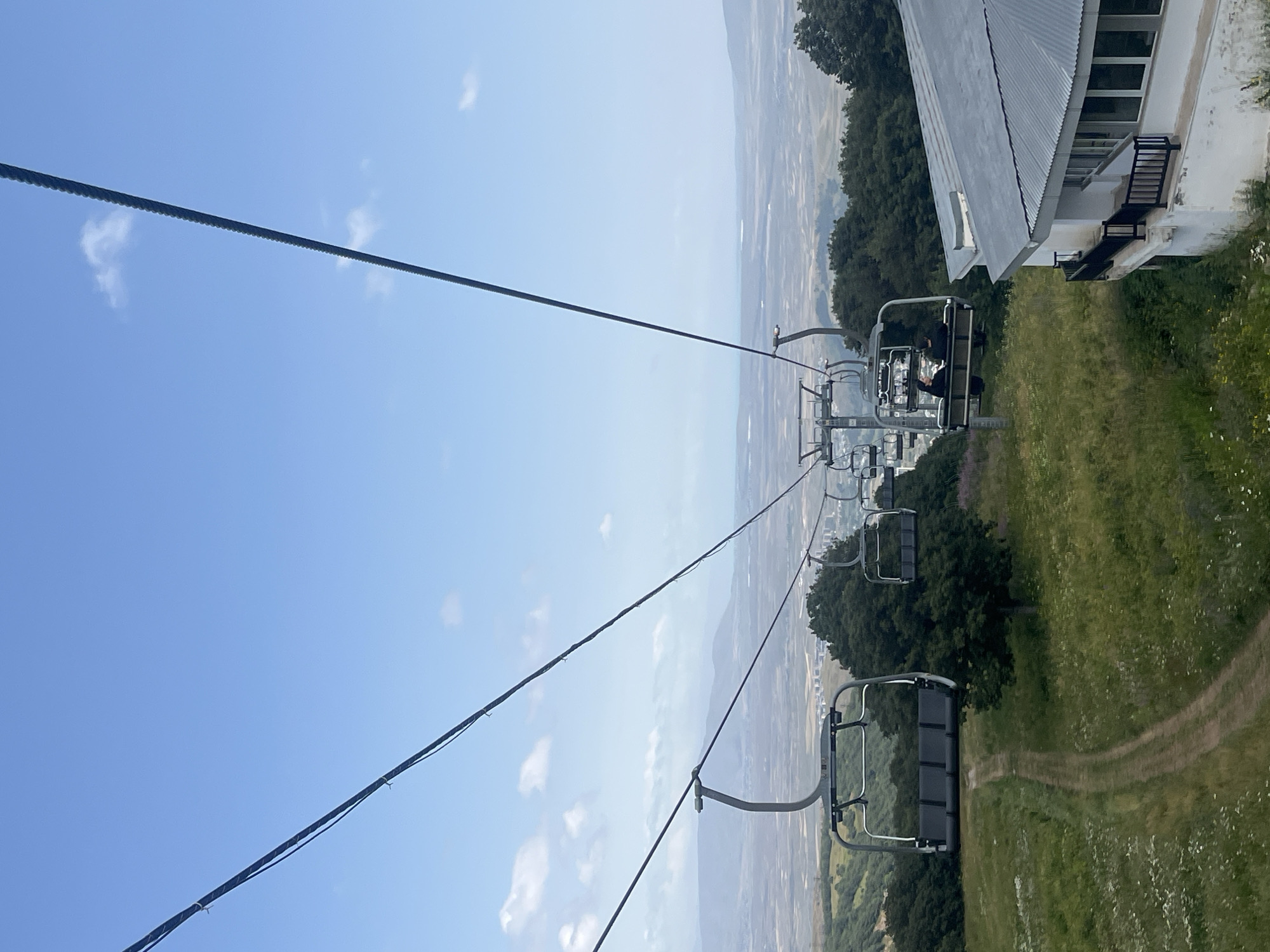 An outdoor, medium shot captures a ski lift moving upwards on a grassy hillside with a view of the city of Tsakhkadzor, Armenia. In the foreground, a white building with a grey, slanted roof and large windows is visible on the right. The ski lift consists of multiple chairs suspended from a thick cable. Two chairs are prominent in the mid-ground, one empty and another carrying two passengers wearing dark clothing. The chairs are dark gray with black seats. The hillside is covered in green grass, scattered with white and purple wildflowers, and features a dirt path winding its way up. Dark green trees dot the landscape. In the background, a hazy landscape of rolling hills and a distant, pale city is visible under a bright blue sky with scattered white clouds. The ski lift cable extends upwards and to the right, disappearing into the sky.