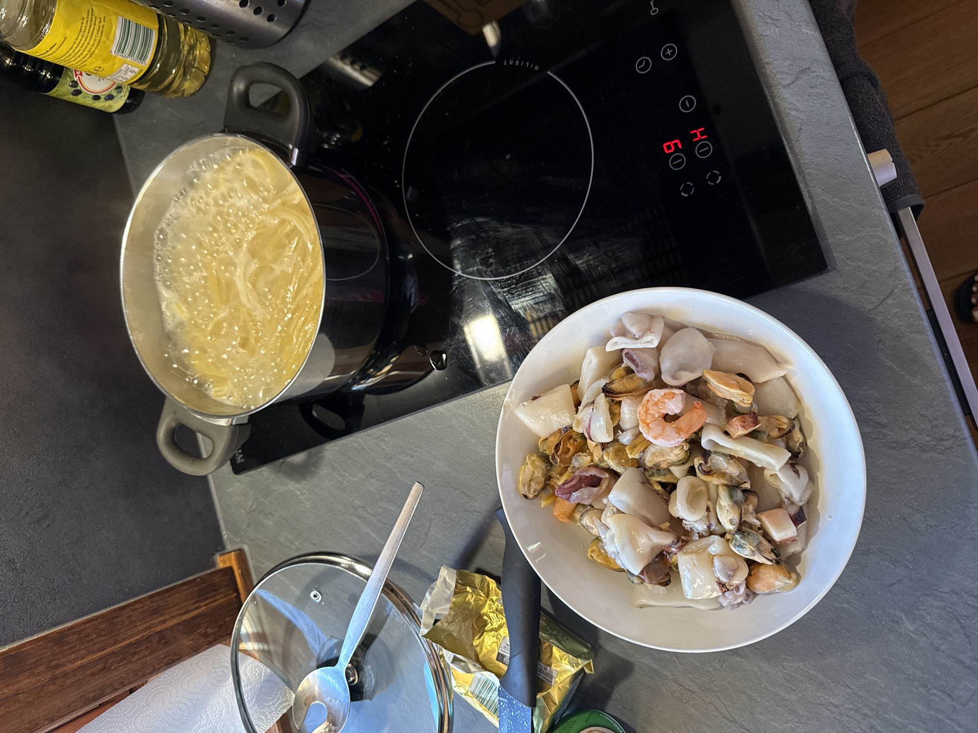 In a kitchen setting, possibly in Breungeshain, Germany, a pot of pasta is boiling on a black induction stovetop. To the left of the stovetop, a bowl of mixed seafood, including mussels, squid, and shrimp, sits on a gray countertop. A spoon rests on a glass lid nearby, next to a roll of paper towels and a packet of butter. The stovetop displays a digital timer showing "09" and "H." Two bottles, one appearing to be olive oil and another dark liquid, are visible in the background on the countertop. The overall scene depicts food preparation in progress.
