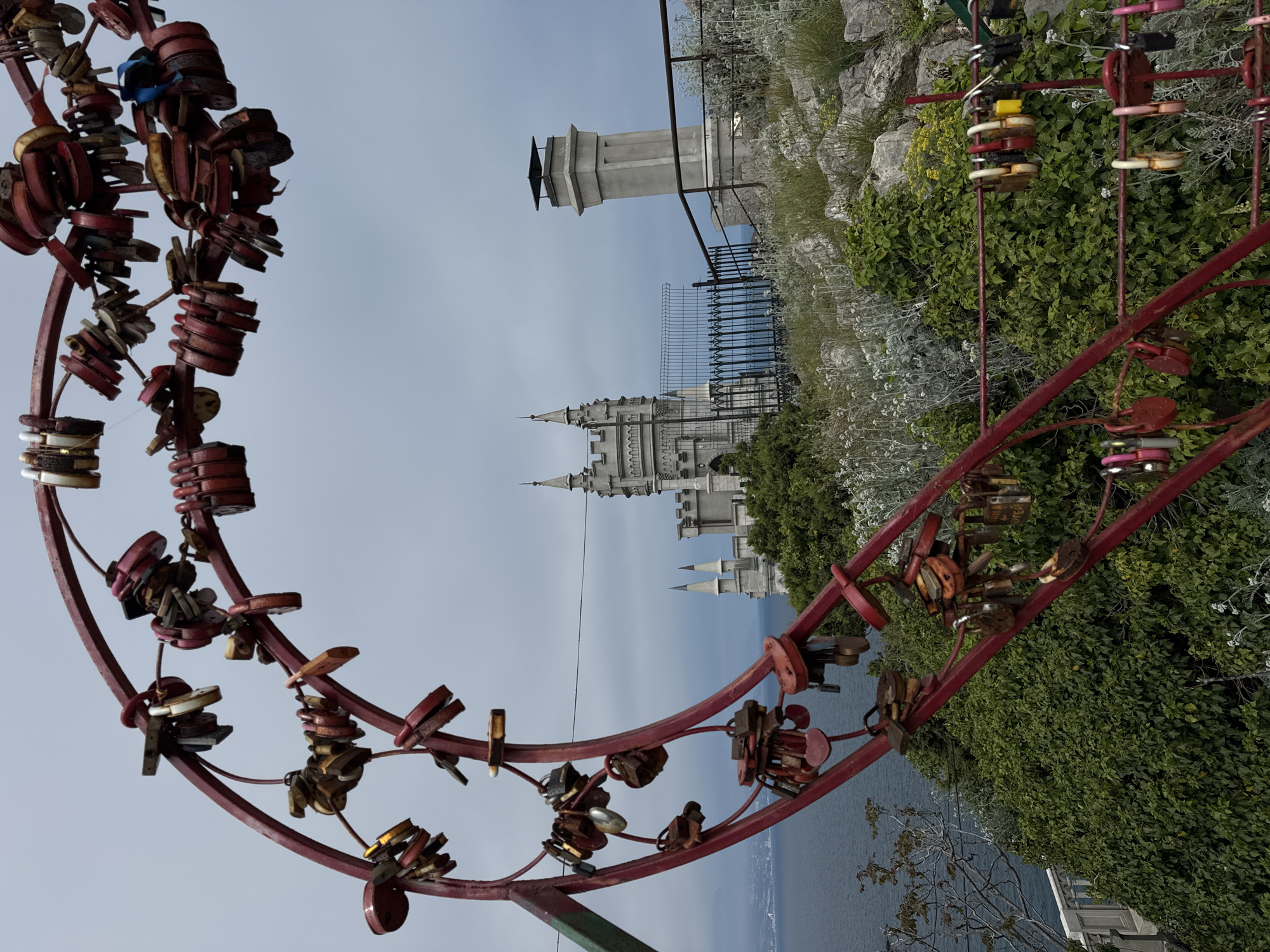 The image shows a red metal structure adorned with numerous padlocks, likely a "love lock" installation. In the background, the Swallow's Nest castle can be seen perched on a cliff overlooking the sea. The vegetation is lush and green, suggesting a coastal environment. The image appears to have been taken in the Гаспринский поселковый совет in Ukraine.