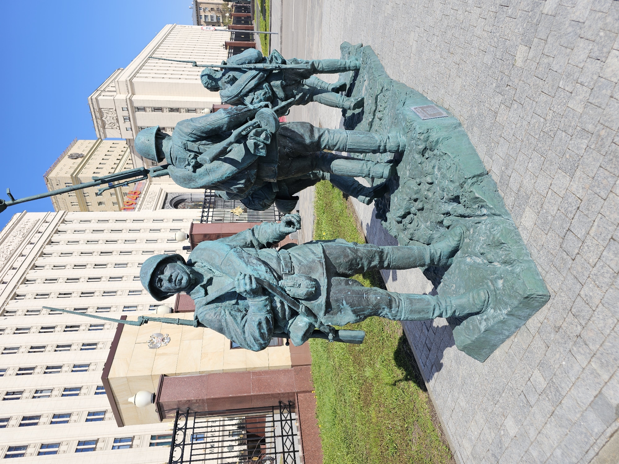 The image shows a bronze monument of three Soviet soldiers located in Moscow, Russia. The monument depicts three soldiers advancing with rifles. The foreground features a soldier with a helmet and full gear. Behind him are two more soldiers in similar attire. The base of the monument is roughly textured and sits on a granite stone base. A patch of grass is behind the statue. In the background is the large light-colored building of the Ministry of Defence. The weather is clear with blue sky. The monument is situated on a grey brick paved area.