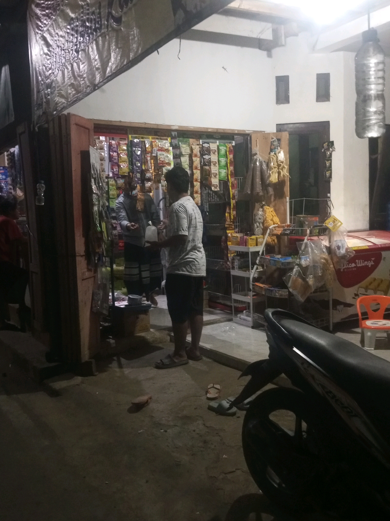 The image depicts a street-level convenience store, likely a 'warung,' in Kalikidang, Indonesia. The store entrance is open, revealing shelves stocked with various packaged goods. Two adult males are engaged in an interaction outside the store. One man, wearing a grey long-sleeved shirt and patterned sarong, appears to be making a transaction or conversation with another man standing to his right. This second man is wearing a light-colored short-sleeved t-shirt and dark shorts, and is holding a clear plastic bottle. The store's facade is adorned with a large, silver banner with text that is difficult to decipher due to the lighting and image quality. Adjacent to the store, a motorcycle is parked, partially obscuring the view of the street. In the background, an upright freezer with branding is visible, along with a small orange plastic chair. The lighting suggests it is nighttime or a dimly lit area. The ground surface appears to be concrete or asphalt.