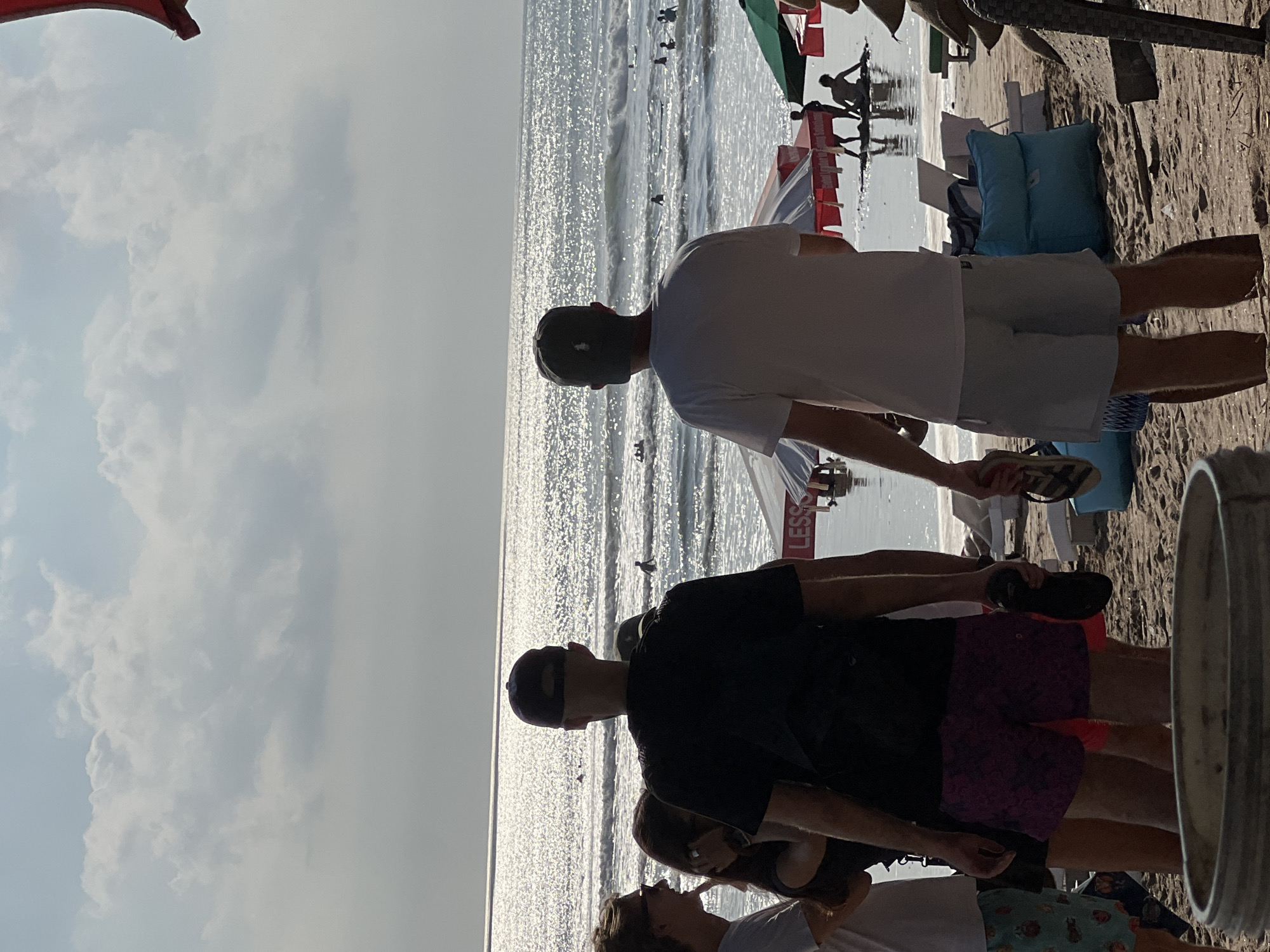 In Kuta, Indonesia, two men stand on a sandy beach, facing the ocean, with other beachgoers in the background. The sea is choppy with gentle waves and several surfers are visible in the water. Beach umbrellas and lounge chairs are scattered on the sand, suggesting a public beach area. The sky is partly cloudy, with the sun casting a bright reflection on the water's surface. The overall atmosphere appears to be a typical day at the beach.