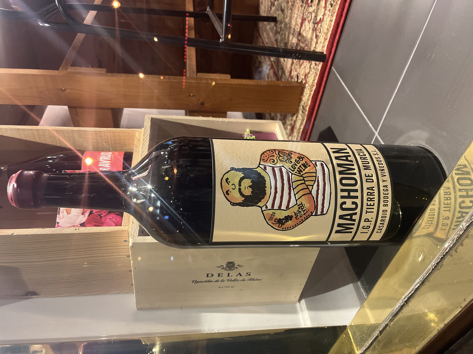 The image depicts a prominently displayed, dark-bodied wine bottle with a red wax-sealed cork, positioned in the foreground of an interior commercial space in Varna, Bulgaria. The bottle's beige label features a stylized illustration of a muscular, bearded man wearing a striped shirt and armbands, with tattoos on his arms, one indicating "2020" and another stating "LOVE & WINE". The label text identifies the product as "MACHOMAN," from "I.G.P. TIERRA DE MURCIA" by "CASAROJO BODEGA Y VIÑEDOS S.L." The bottle rests on a dark gray tiled surface, with a tarnished brass-colored reflective strip immediately in front of it, showing a partial reflection of the label. In the background, various elements of a wine retail or hospitality environment are visible: light-colored wooden wine crates, one clearly marked "DELAS Vignobles de la Vallée du Rhône 6 x 750 ml", and another bottle with a partially visible red label reading "FAVRUD". Wooden shelving and structural beams form part of the background architecture. The floor in the mid-ground is covered by a patterned red rug, and a black metal leg, consistent with a chair or bar stool, is visible on the right. The scene is illuminated by multiple warm, diffuse light sources, creating a bokeh effect. No individuals are present in the frame.