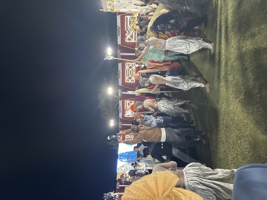 A nighttime outdoor scene shows people dressed in traditional Indian attire, some with body paint. Many are lying on the ground, while others are standing, holding cameras. A decorated structure with lights is behind them. The scene appears to be a cultural performance or procession. The lighting is dim, with several bright spotlights illuminating the event. The background is mostly dark sky.
