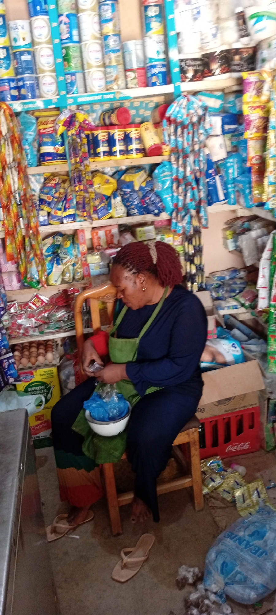 This image captures the interior of a small, well-stocked shop, likely a local convenience store, in Gayawa, Nigeria. The scene is dominated by a woman, appearing to be middle-aged, with red dreadlocks tied in a ponytail. She is wearing a dark blue long-sleeved top and a green apron, and is seated on a simple wooden stool.
The woman is actively engaged, looking down at her hands, where she holds a small item over a white bowl that contains a blue plastic bag. This suggests she might be weighing or packaging goods for a customer. Her light-colored flip-flops are visible on the concrete floor beside her.
The shop itself is densely packed with various products arranged on wooden shelves. The top shelves are filled with numerous tins of milk powder, identifiable by brands such as "NAN" and "PUREMILK." A shelf divider prominently displays the text "BREMFAST TIME." Lower shelves showcase an array of packaged items, including yellow bags of "Chelsea" sugar, "SUPREME NOODLES" with "TASTY CHICKEN" flavor, and other household goods. Bundles of sachets hang from strings, adding to the cluttered yet functional appearance.
On the left side of the frame, a tray of eggs is visible, along with a package labeled "CHERIE 20g." The floor on the right features a red "Coca-Cola" crate and a large blue plastic bag with the brand name "JEANIE," surrounded by some scattered wrappers. The overall impression is that of a bustling, essential local business. The lighting is ambient, with no distinct time of day or weather indicated.