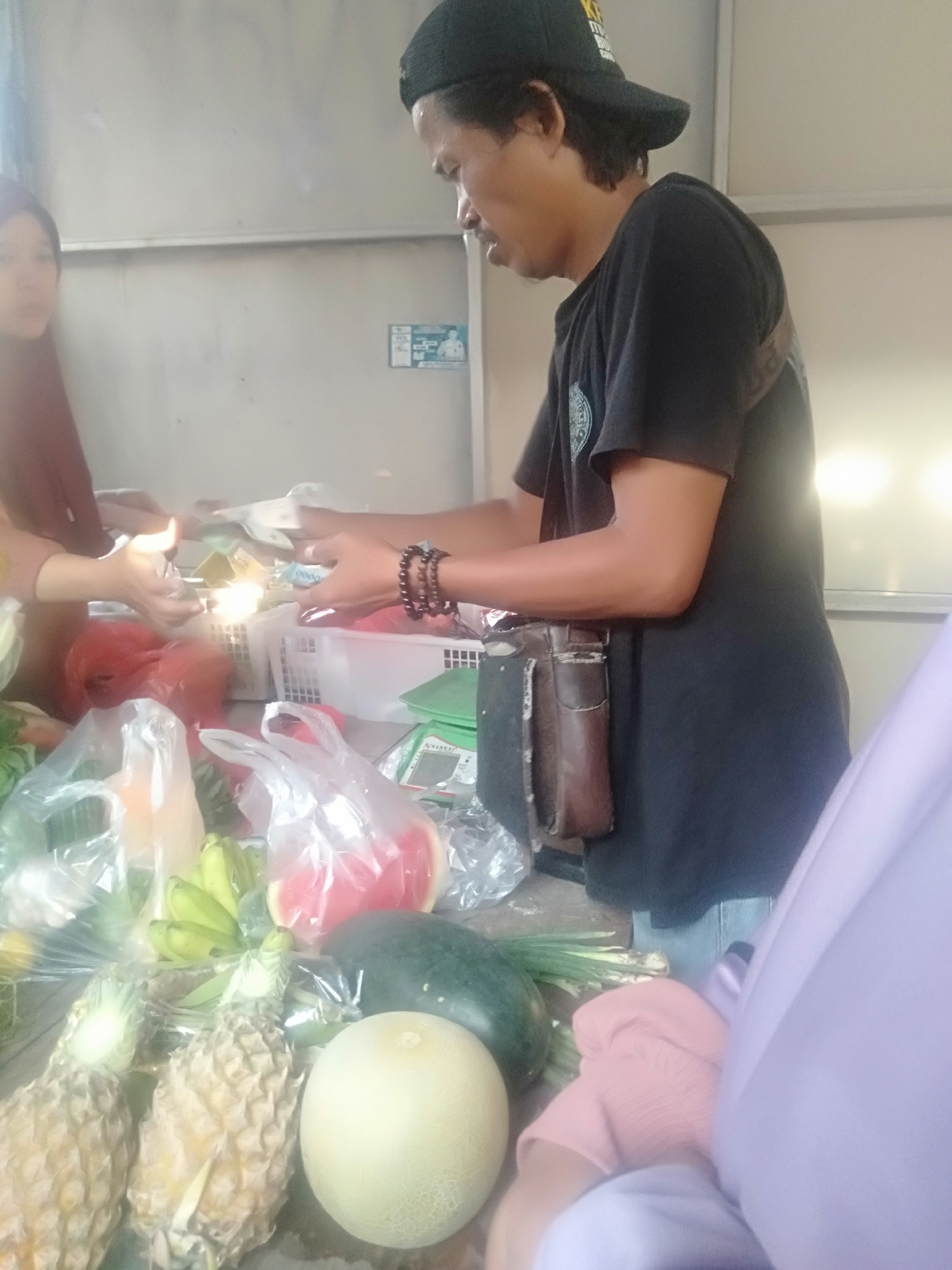 The image depicts a transaction at what appears to be an outdoor market stall in Sukadami, Indonesia. A man wearing a black t-shirt and a black baseball cap is handing money to another person whose hands are visible from the left side of the frame. The man has a brown pouch attached to his belt. In the foreground, a variety of fruits are displayed, including pineapples, bananas, a watermelon, and a melon, some of which are wrapped in plastic. Other items, such as plastic bags and a white basket, are also visible. The background consists of a plain wall with a poster and some shelving. The lighting suggests it is daytime.