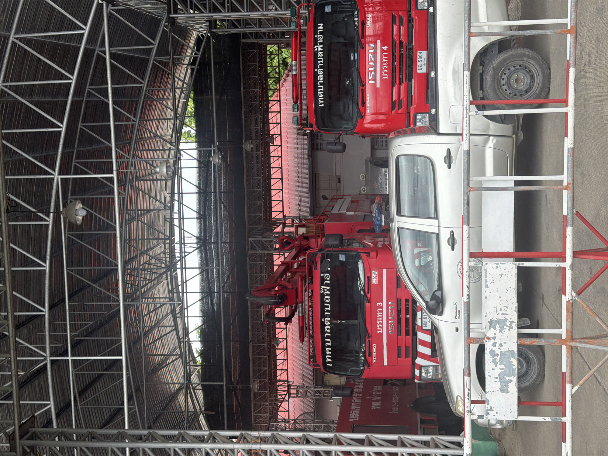 The image shows a fire station in the city of Chang Phueak, Thailand. The fire station has a metal roof structure supported by metal poles. In front of the station, there are three vehicles: two red fire trucks and one silver pickup truck. The fire trucks are parked side by side, and the pickup truck is parked in front of them. The fire trucks have the words "เทศบาลตำบลฟ้าฮ่าม" written on them in Thai script, which translates to "Tha Phae Sub-district Municipality". One fire truck also has the text "บรรเทา 3" on it, meaning "Relief 3". The pickup truck has the text "HILUX D4D" on its side. The scene appears to be calm and orderly, with no visible human activity. The environment is outdoor, with trees visible in the background.