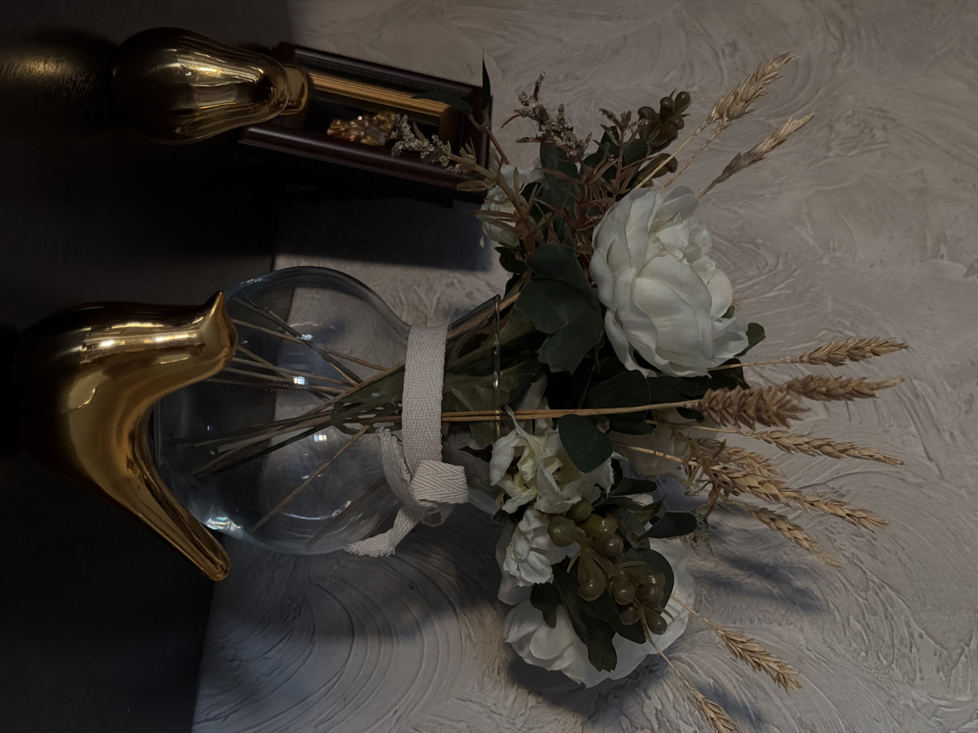 The image depicts a floral arrangement in a glass vase with a gold stand, secured with a white cloth ribbon. The arrangement includes white flowers, green leaves, wheat stalks, and berries. The vase is positioned against a textured wall, with parts of a gold-colored object and a dark wooden frame visible in the background. The setting appears to be indoors. The city of Rossiyskiy, Russia, is mentioned but cannot be confirmed from the image.