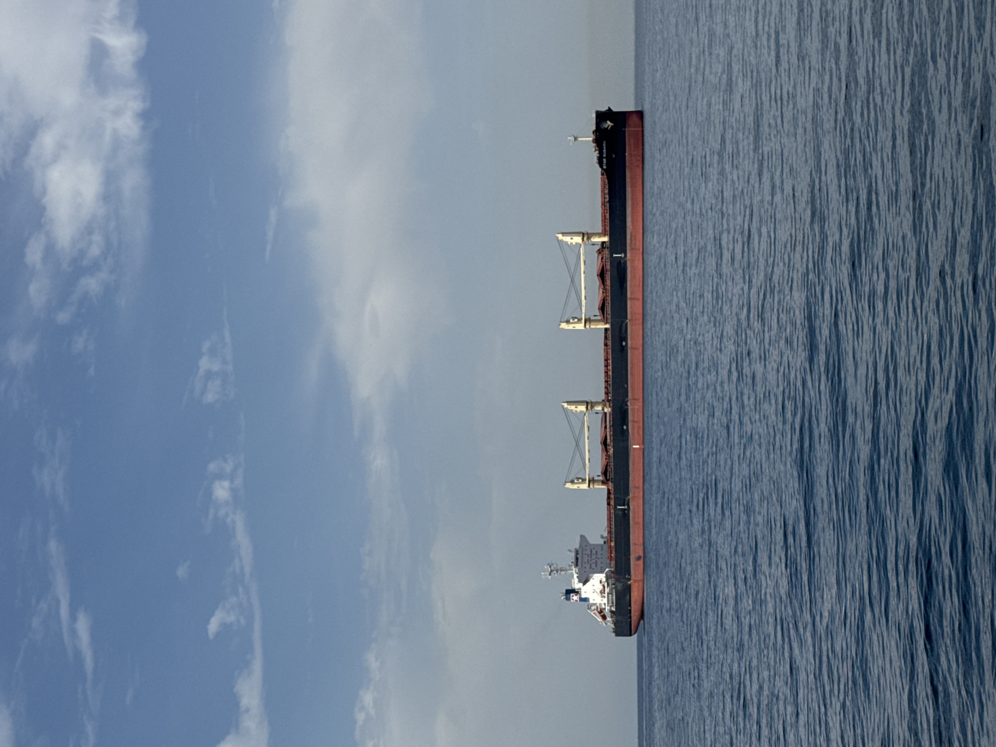 An offshore freighter with a black hull and a red waterline is depicted moving across calm blue water under a partly cloudy sky. The freighter has a white superstructure with a tall red, white, and blue smokestack. The vessel has several large cargo holds with visible cranes. The name "STAR SUBARU" is visible on the side of the hull near the stern. The scene suggests a location within the port of Marina di Ravenna, Italy, given the type of vessel and its typical presence in such industrial port areas.